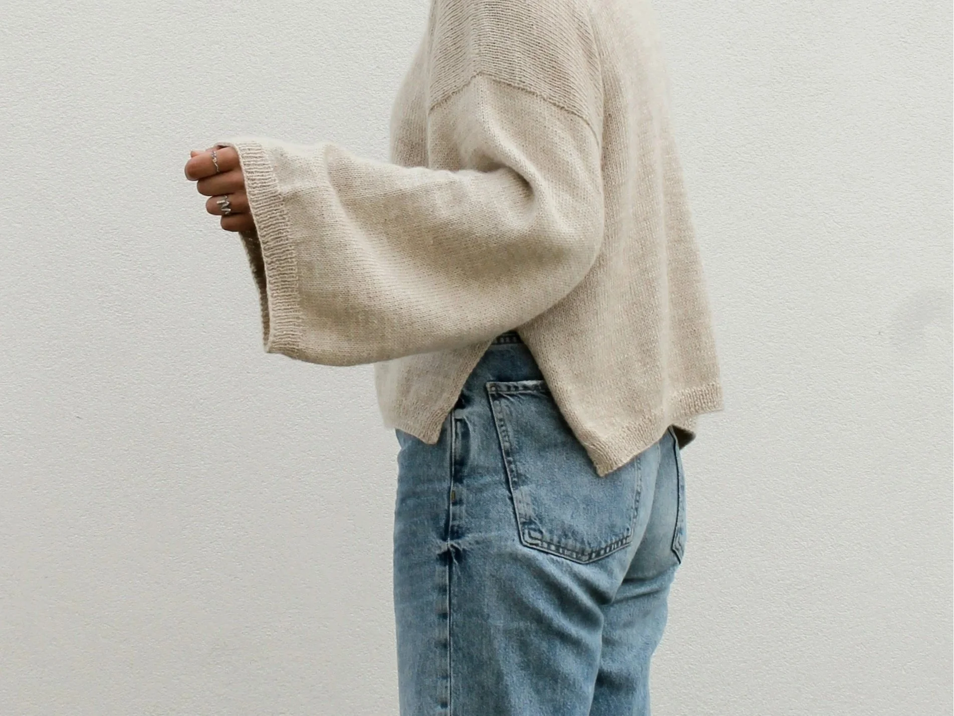Person wearing a beige oversized sweater and light blue jeans standing against a plain white wall.