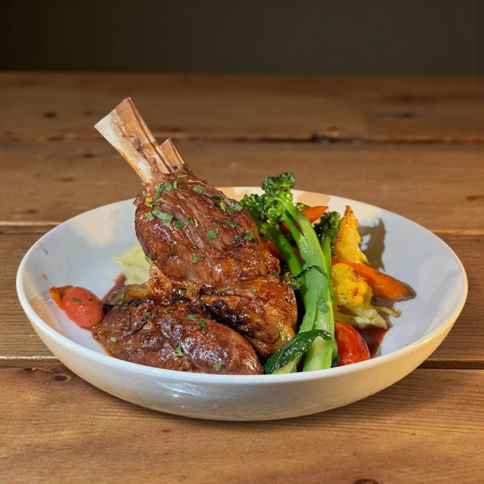 A white bowl of cooked lamb shank with vegetables including broccoli, carrots, and potatoes on a wooden table.