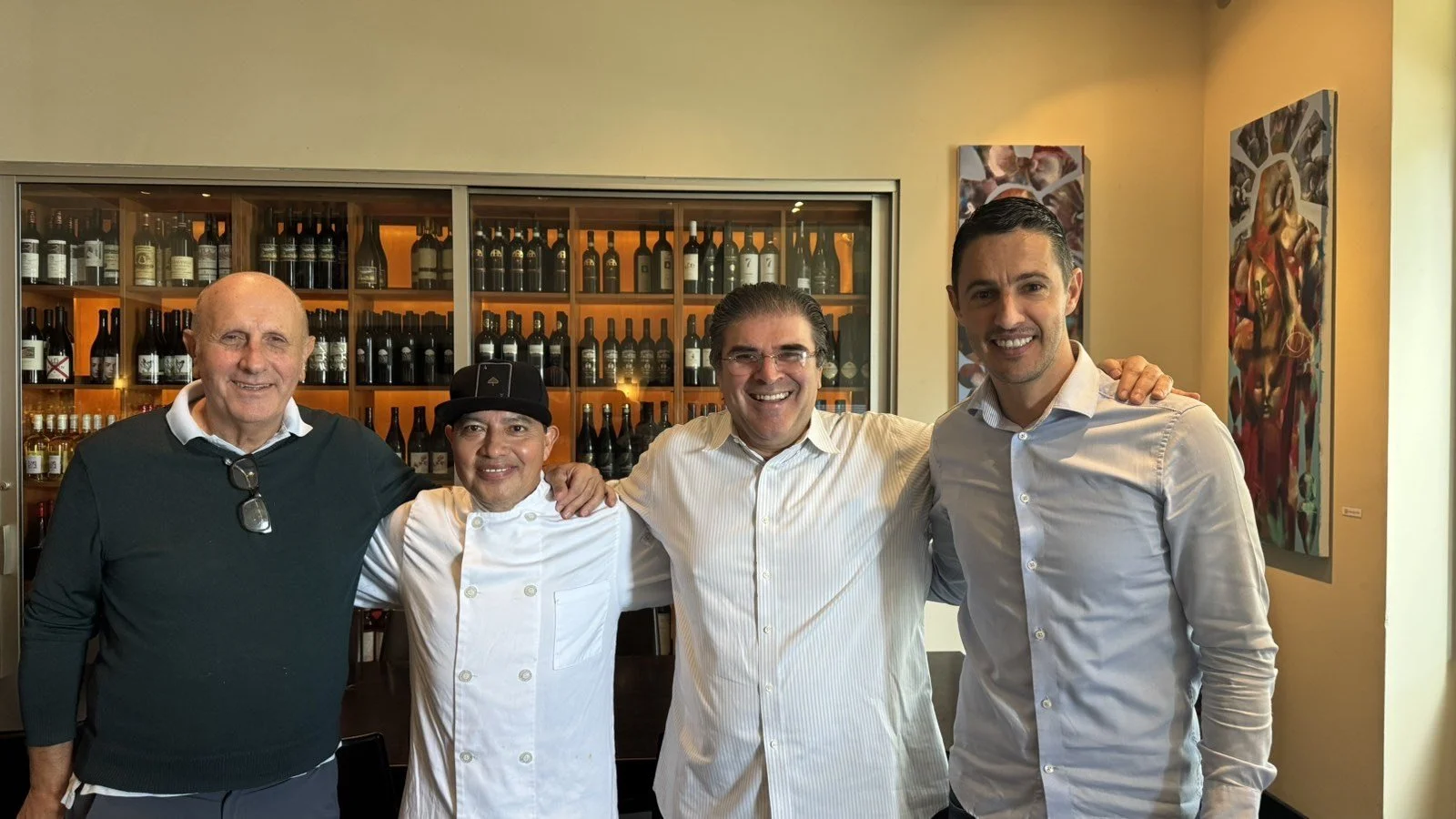 Four men standing together indoors, smiling at the camera. The background features a wine display and colorful artwork on the walls.