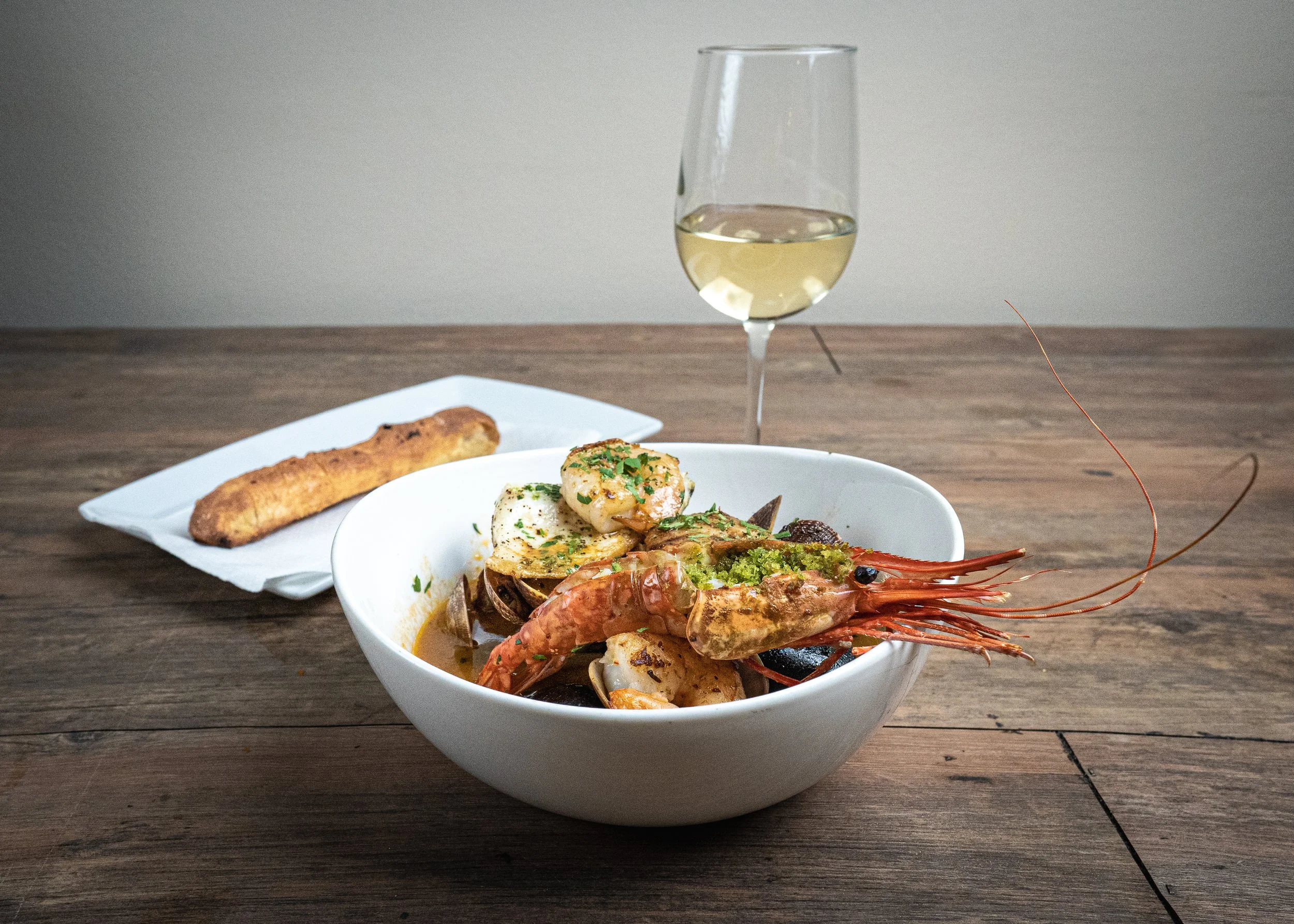 Seafood dish with lobster, scallops, and other shellfish in a white bowl, served with a piece of bread and a glass of white wine on a wooden table.