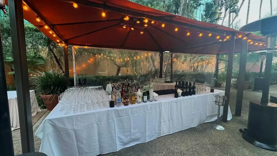 Outdoor bar setup with a white tablecloth, bottles of alcohol, glasses, and string lights under a large red canopy.