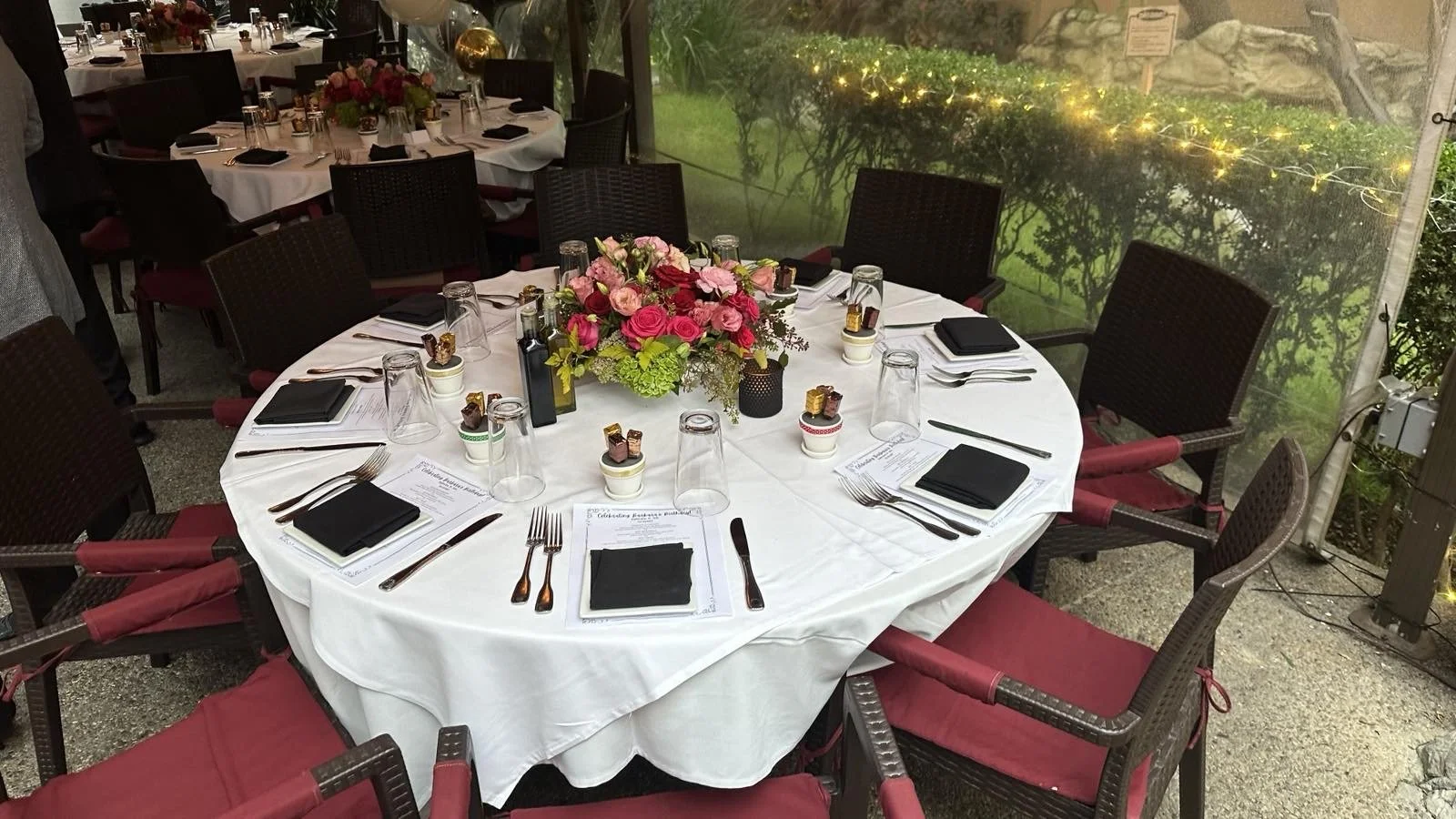 Round table set for a formal event with a white tablecloth, black napkins, glasses, silverware, and a floral centerpiece of pink, red, and green flowers, situated outdoors with a grassy area and decorative lights in the background.