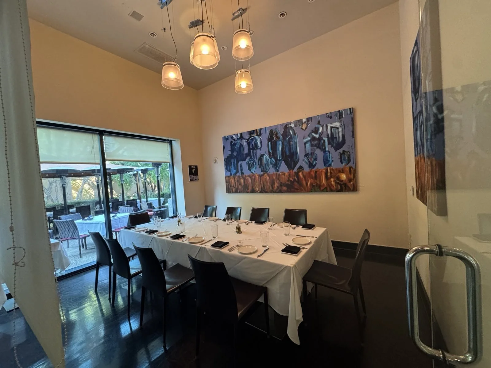 A dining room with a rectangular table set for a meal, featuring white tablecloths, black plates, silverware, and glasses. There are black chairs around the table. The room has a large window/door that opens to an outdoor patio with umbrellas. Modern ceiling light fixtures hang from the ceiling. The wall has colorful abstract artwork.