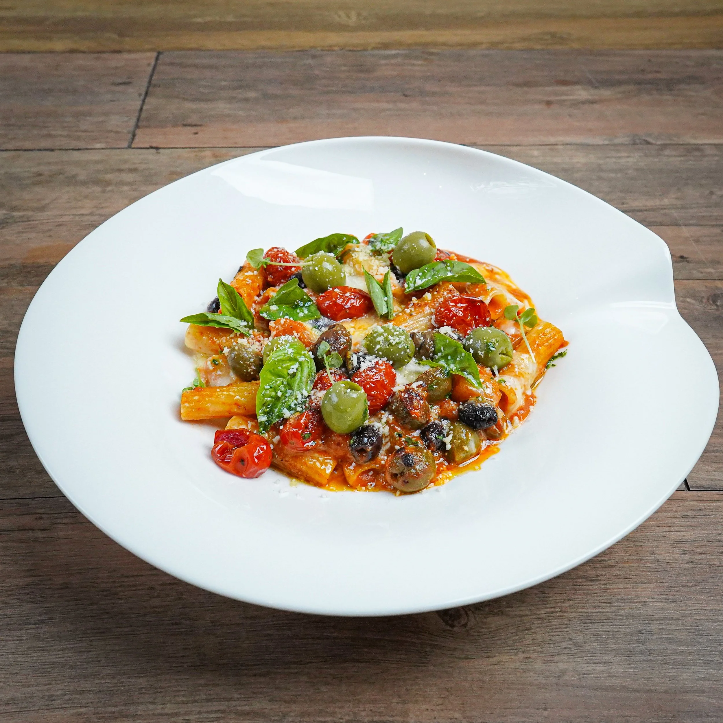 Plate of pasta with olive, cherry tomatoes, basil leaves, and grated cheese on a wooden table.