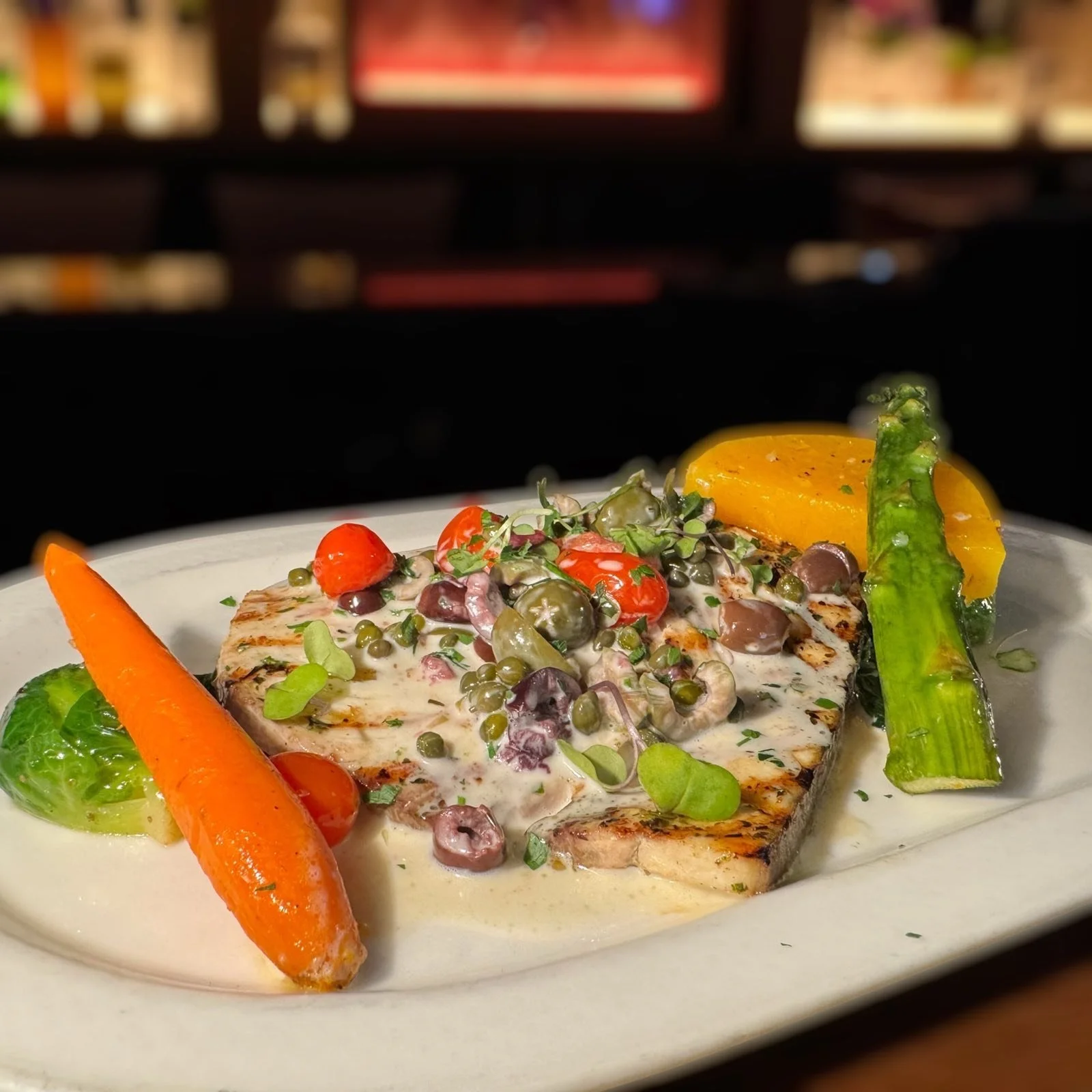 Grilled fish fillet topped with creamy sauce, cherry tomatoes, capers, and microgreens, served with roasted carrot, zucchini, and yellow squash on white plate.