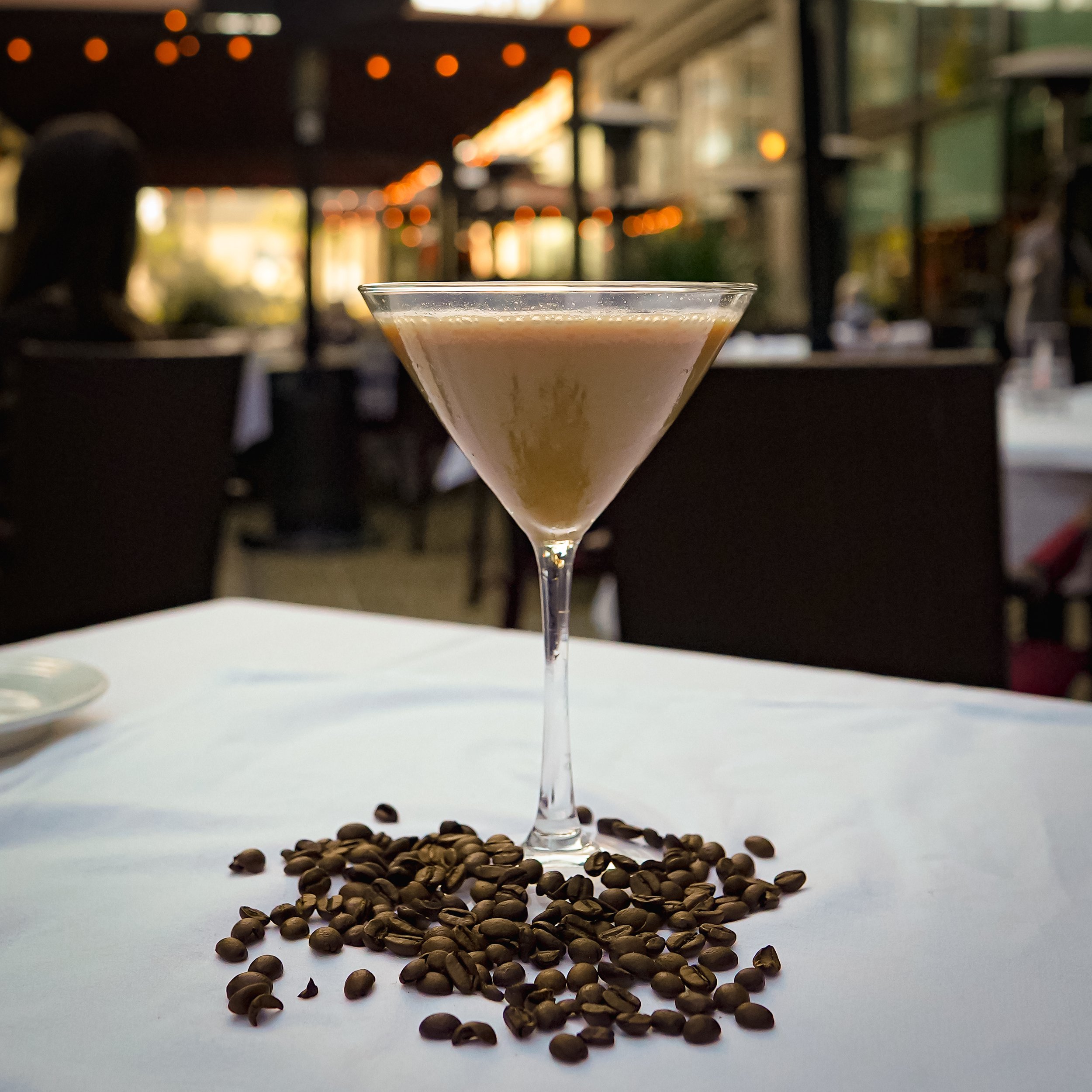 Martini glass with a creamy beverage on white tablecloth, surrounded by coffee beans, in an outdoor restaurant setting with blurred background and warm string lights.