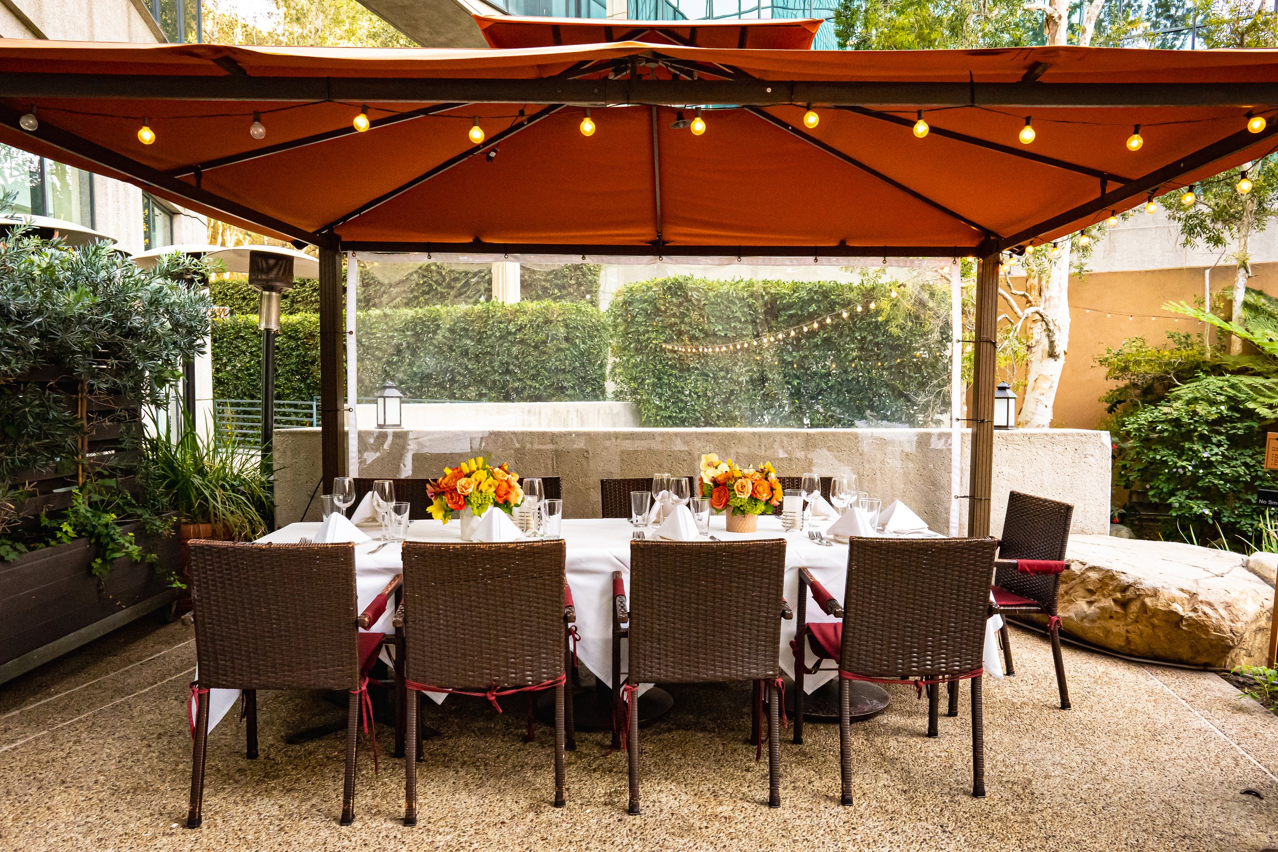 Outdoor dining setup with a long white tablecloth, floral centerpieces, and eight chairs, under a large orange umbrella with string lights, surrounded by greenery and a concrete wall.