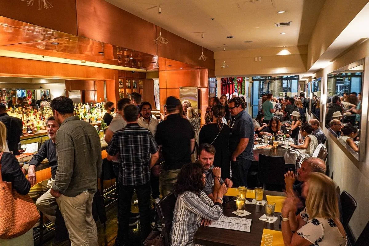 A busy restaurant with people sitting at tables and standing at the bar, socializing and enjoying drinks and food.