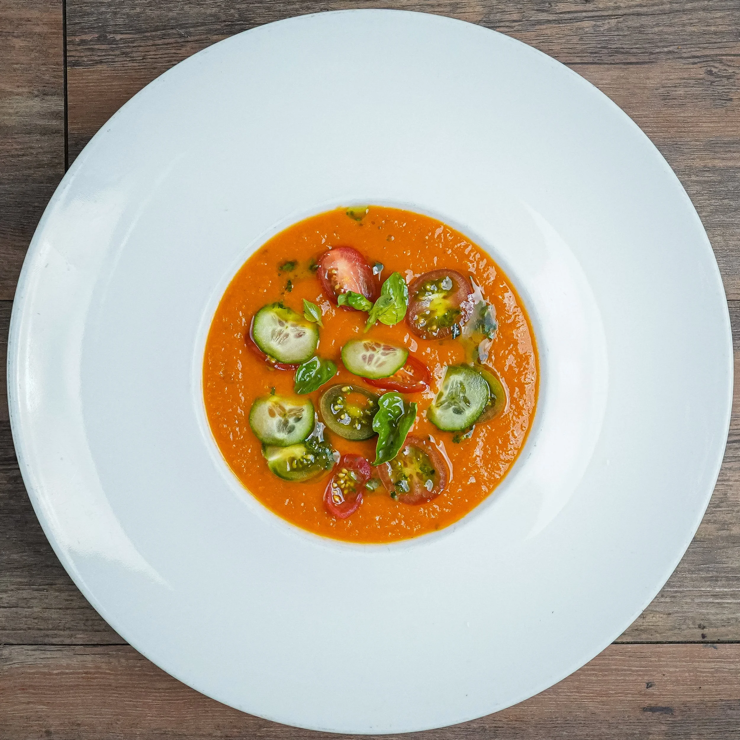 A bowl of tomato-based soup garnished with sliced cucumbers, cherry tomatoes, jalapeños, and basil leaves, served on a white plate on a wooden table.