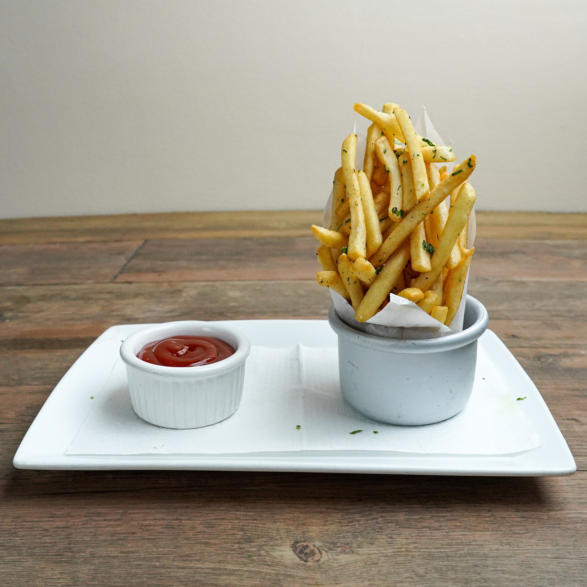 A serving of French fries topped with herbs served in a small white cup, accompanied by a small ramekin of ketchup on a white rectangular plate.