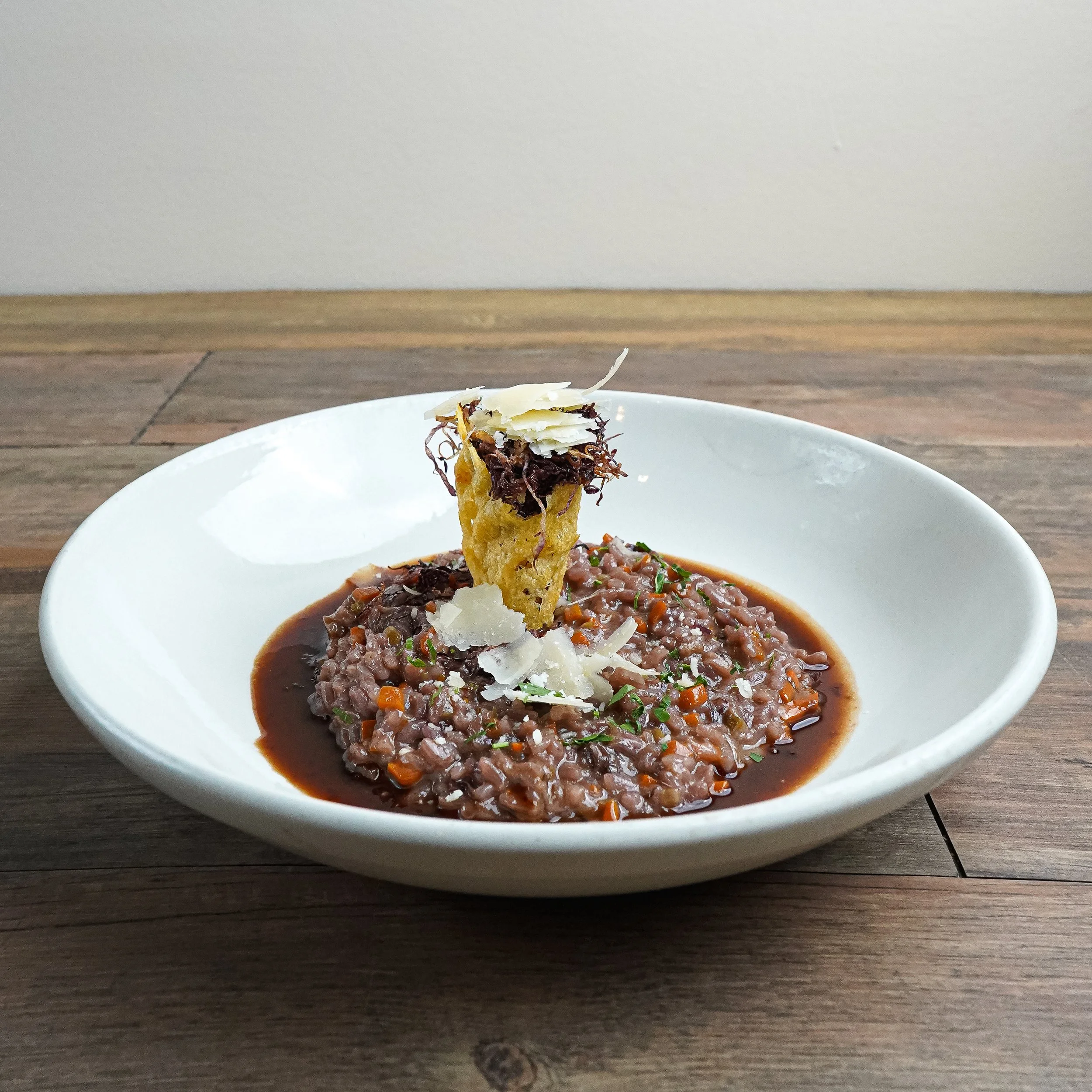 Bowl of beef stew with vegetables, topped with a fried onion ring, shaved cheese, and herbs, on a wooden table.