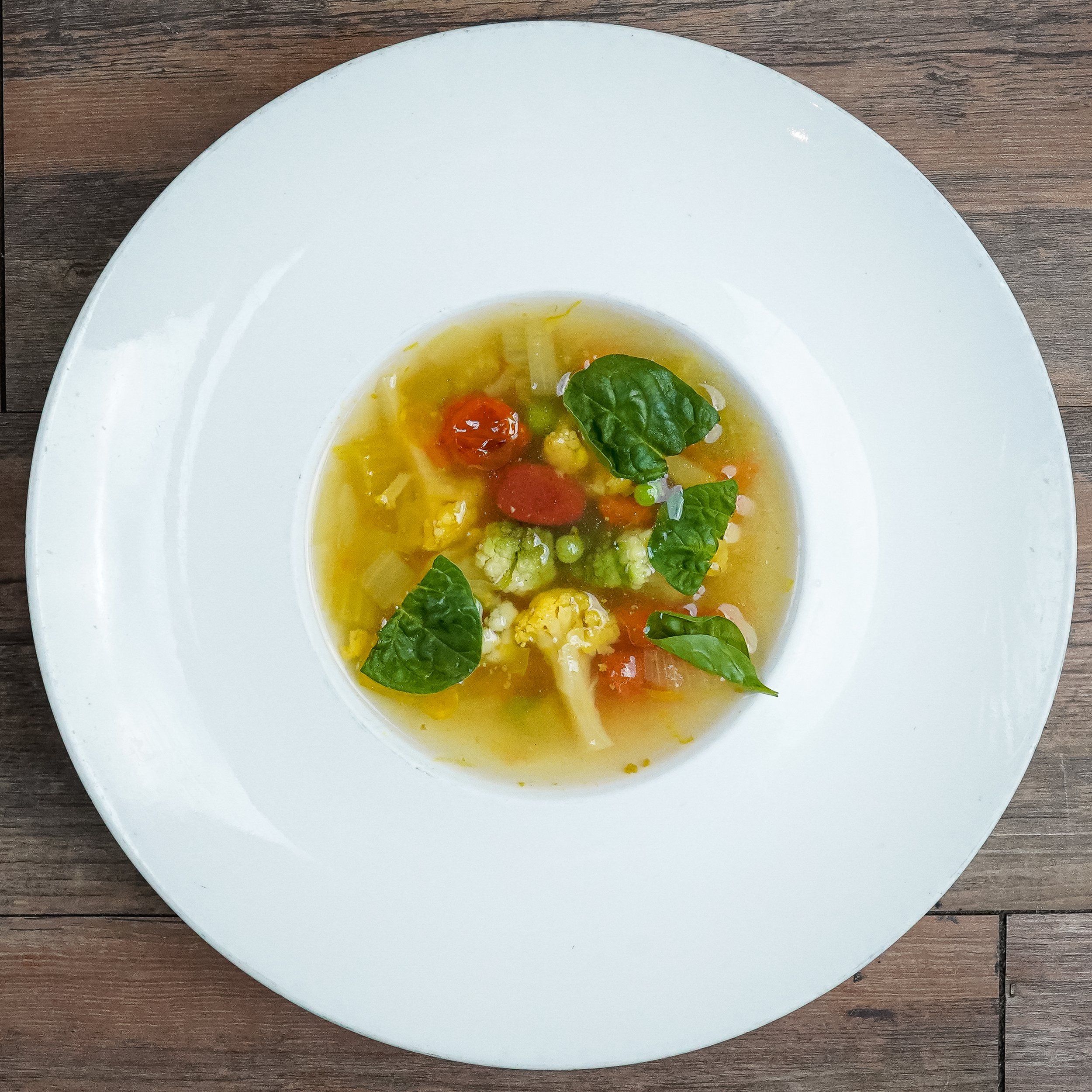 Bowl of vegetable soup with cauliflower, cherry tomatoes, peas, carrots, celery, and fresh basil leaves on top