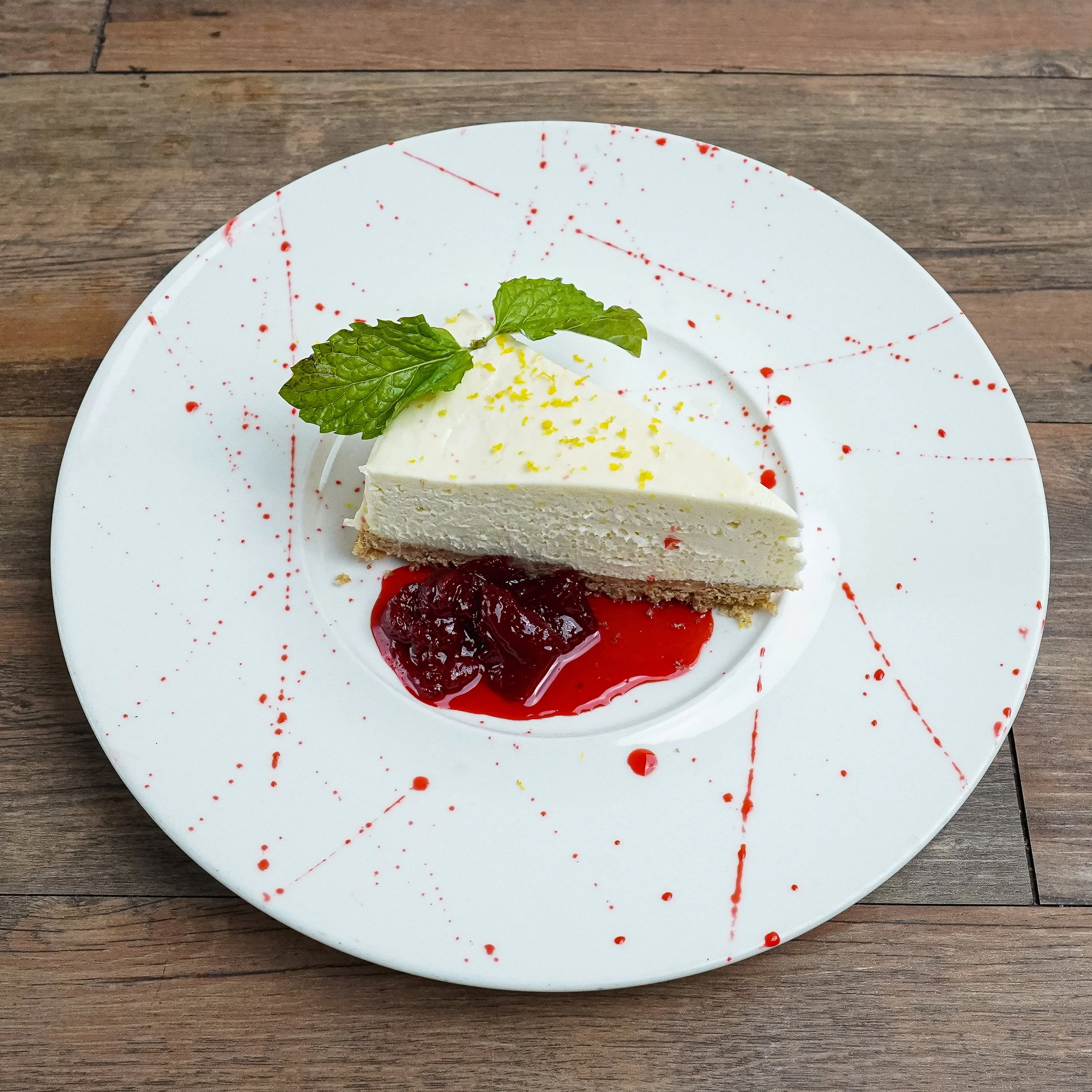 Slice of cheesecake with lemon zest and mint leaves on top, served with berry compote on a white plate with red splatter design, placed on a wooden surface.