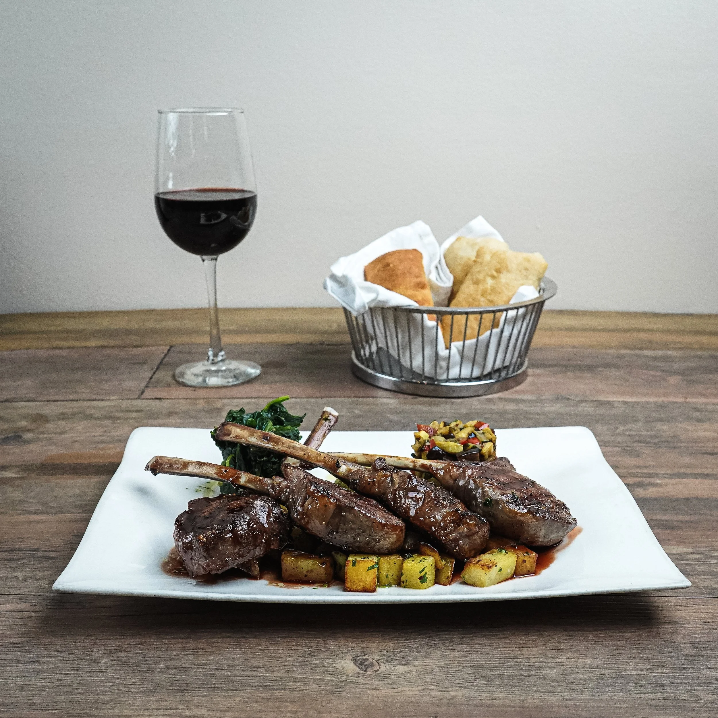 A plate of lamb chops with diced potatoes, spinach, and a side of chopped vegetable salad, accompanied by a glass of red wine and a basket of fried bread on a wooden table.