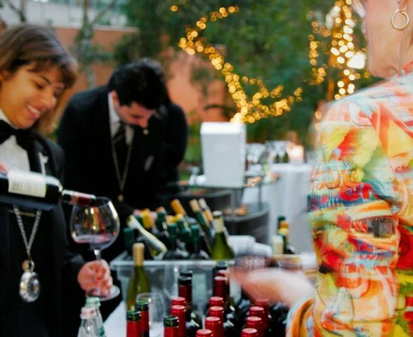 People at a wine tasting event with bottles of wine on display and string lights in the background. Just one of the things you can plan at IL Moro for future events