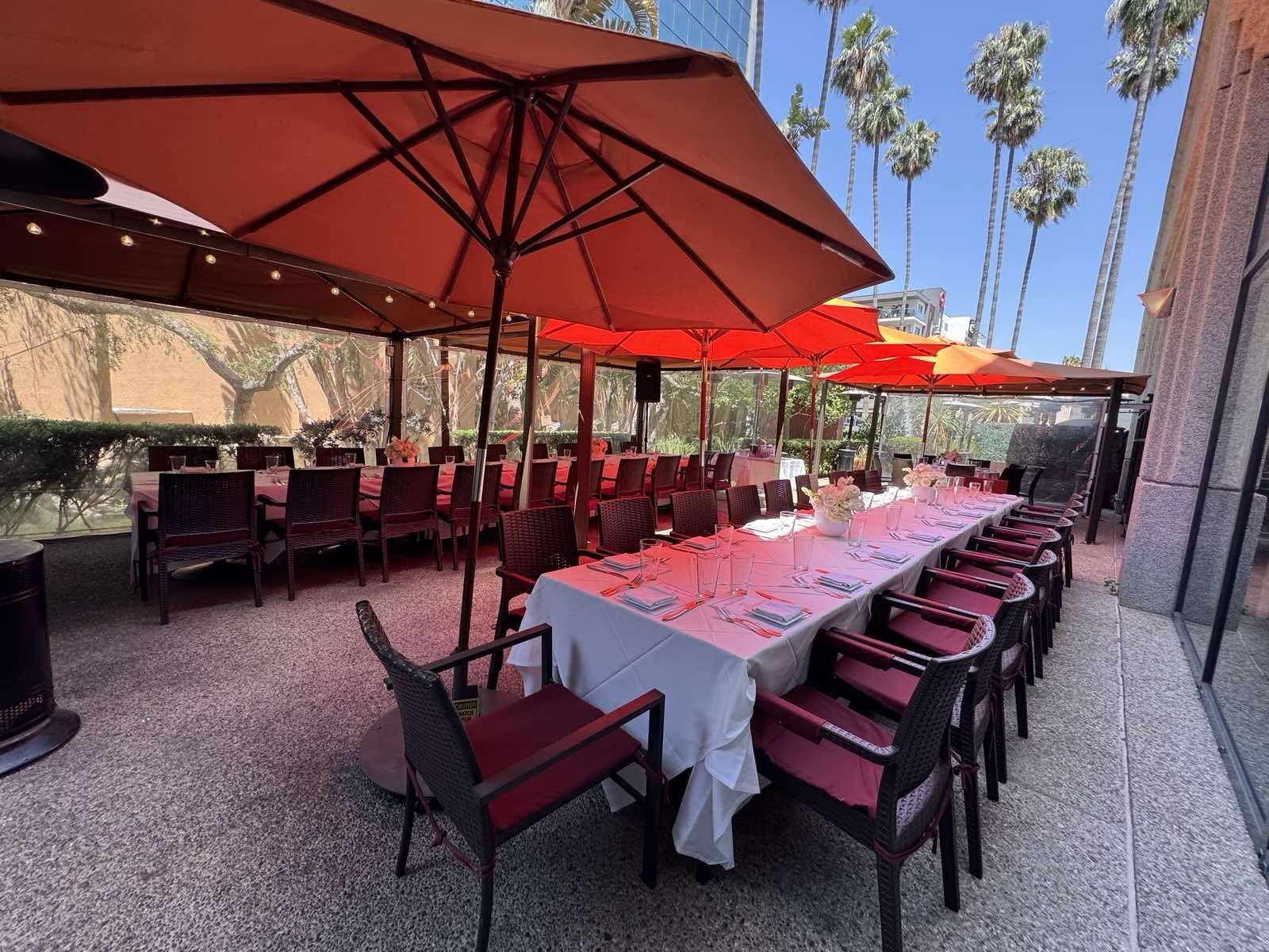 Outdoor dining setup with long table covered with a white tablecloth, adorned with floral centerpieces, set for a meal with glassware and cutlery. Large red umbrellas are providing shade, and there are additional chairs arranged along the sides. The background shows a sunny day with palm trees and a clear blue sky.