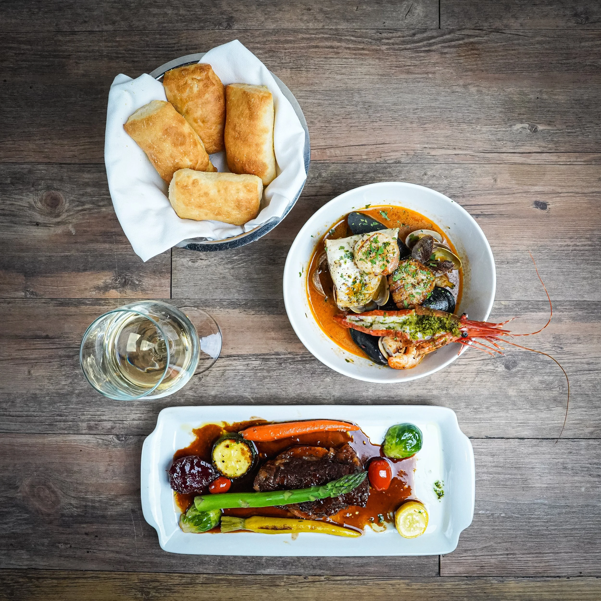 A wooden table with a basket of fried spring rolls, a bowl of seafood stew with shrimp and mussels, a glass of white wine, and a plate of braised meat with vegetables and sauce.