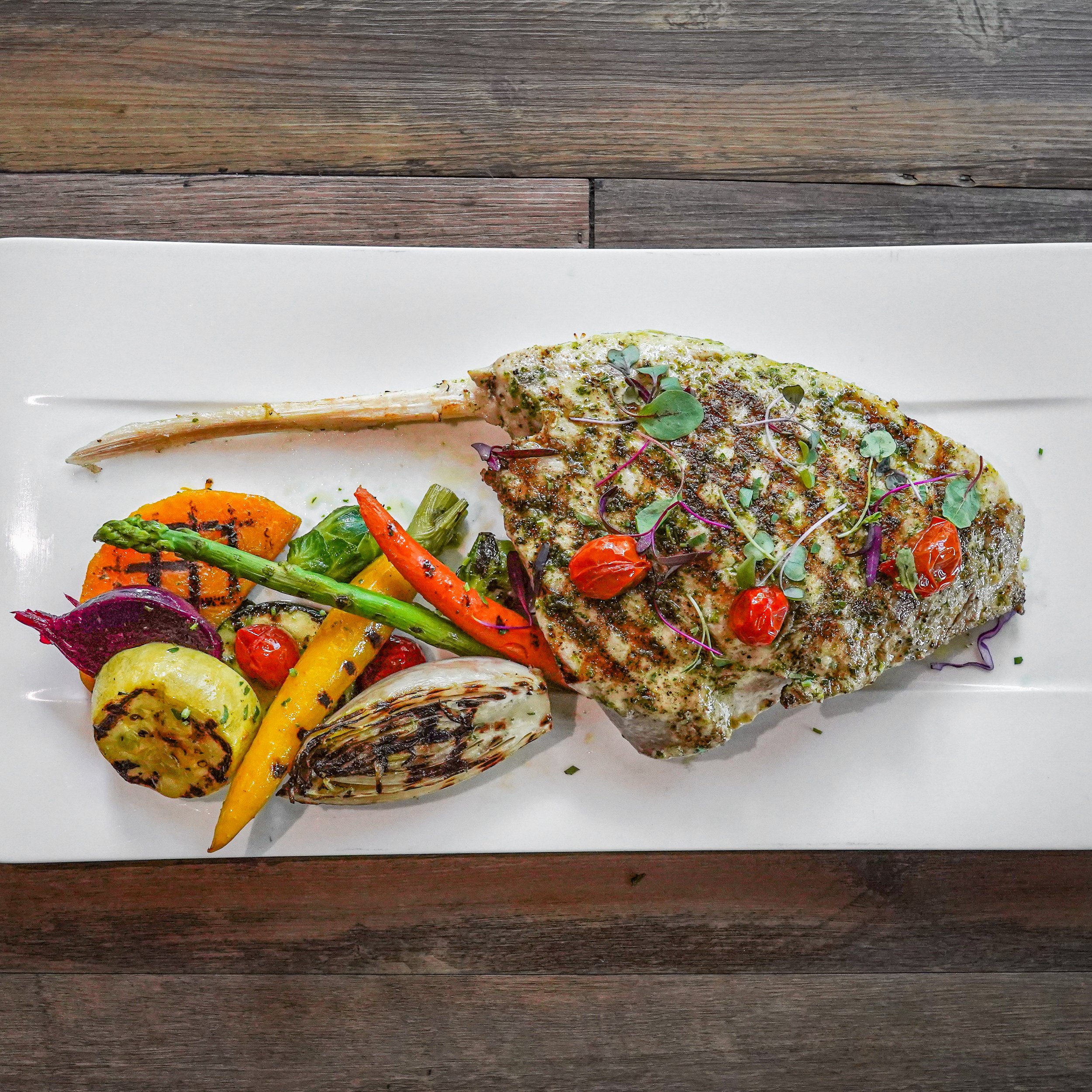 Grilled fish fillet garnished with microgreens and cherry tomatoes, served with grilled vegetables including carrots, zucchini, asparagus, and red onion on a white rectangular plate.