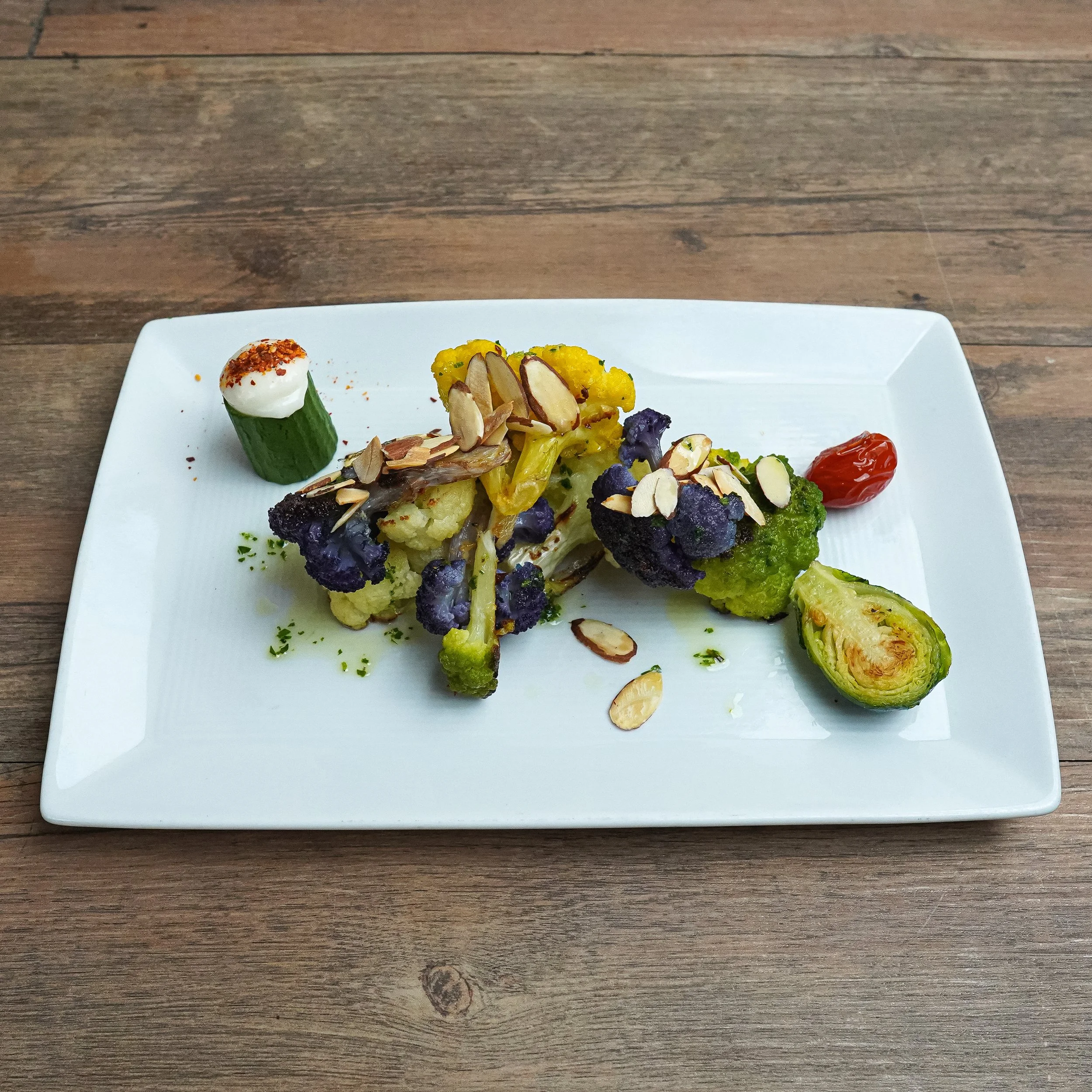 Colorful roasted vegetables garnished with sliced almonds and served with cherry tomato and a piece of stuffed brussels sprout on a white rectangular plate.