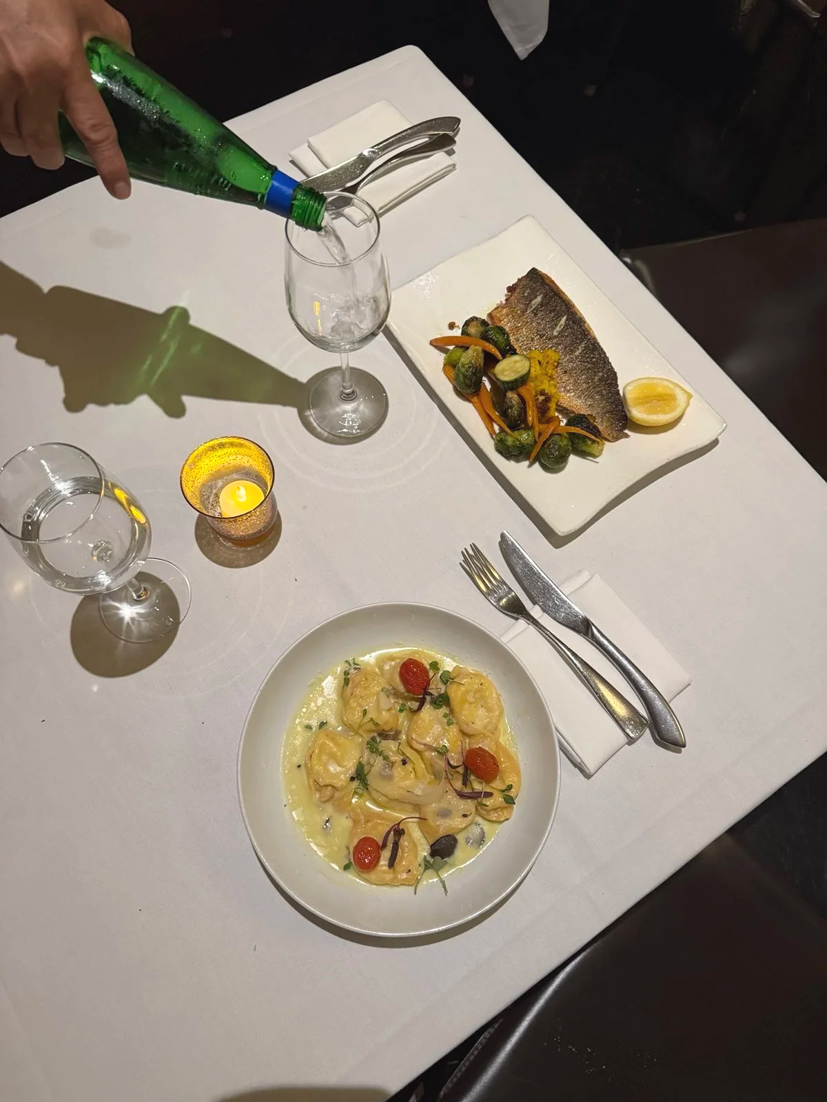 A table with a white tablecloth set with a plate of ravioli in a creamy sauce garnished with herbs and cherry tomatoes, a plate with a grilled fish fillet with vegetables and lemon on a white rectangular plate, an empty wine glass, a glass with water, a hand pouring a green drink into the water glass, a small candle, and silverware wrapped in a napkin.