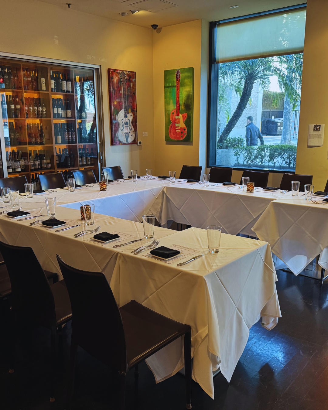 A restaurant dining area with a U-shaped table set with glasses, black napkins, and cutlery. The room has a large window with a view of trees and a person walking outside, and there is wall art of colorful guitars and a wine display cabinet.