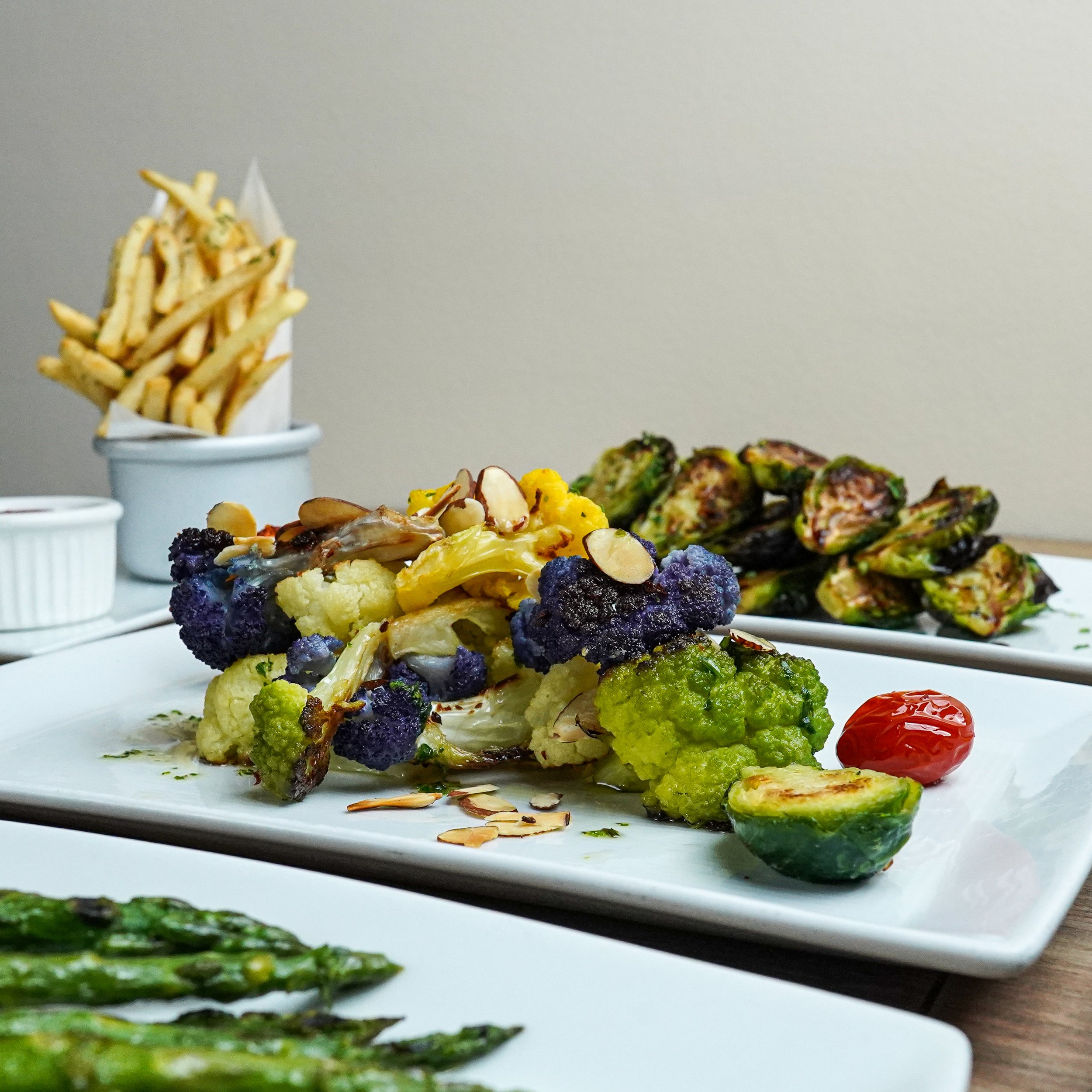 A plate of roasted vegetables including purple and green cauliflower, a cherry tomato, and grilled zucchini, with other plates of grilled asparagus and a side of French fries in the background.