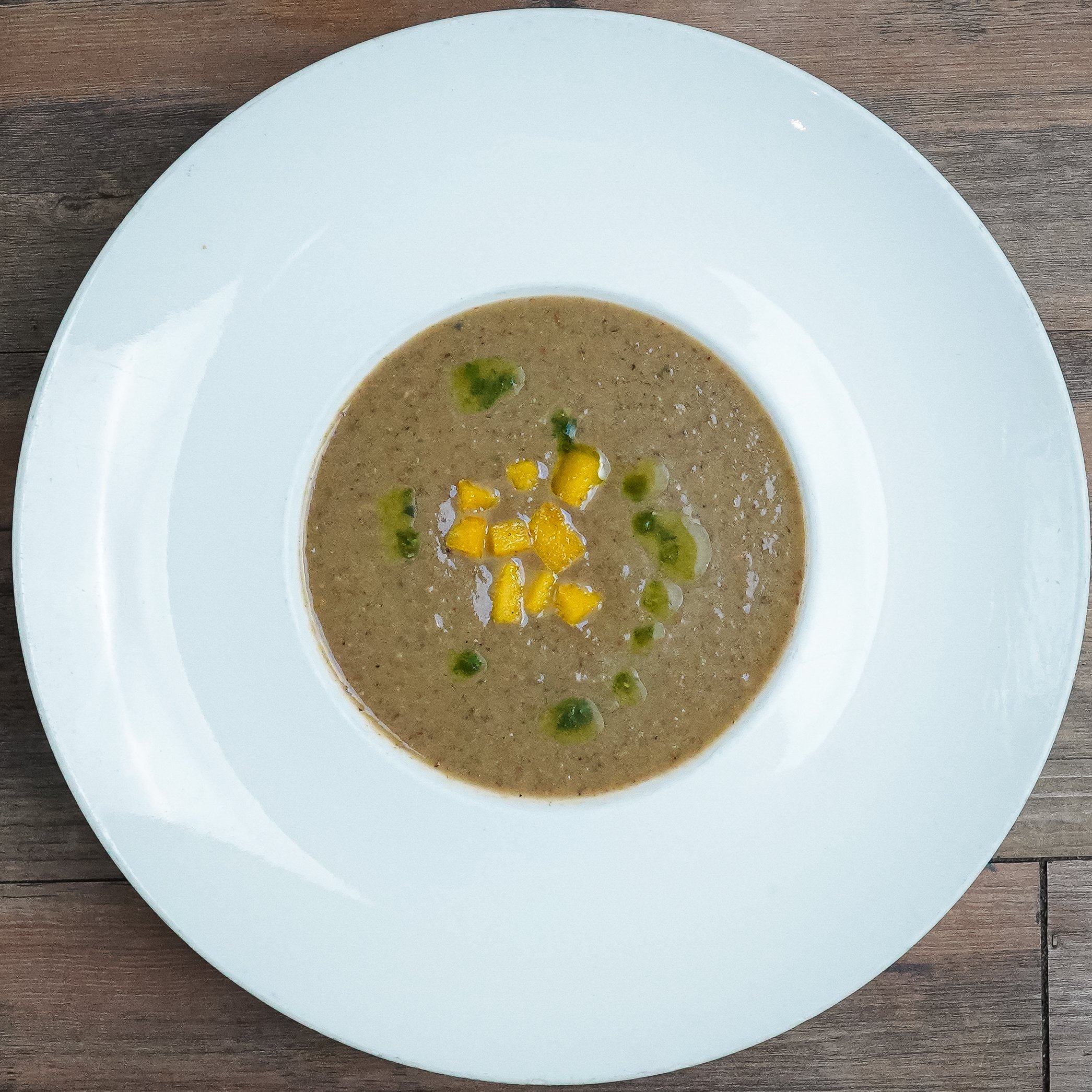 A white plate with a bowl of mushroom soup garnished with chopped green herbs and yellow corn kernels, placed on a wooden table.