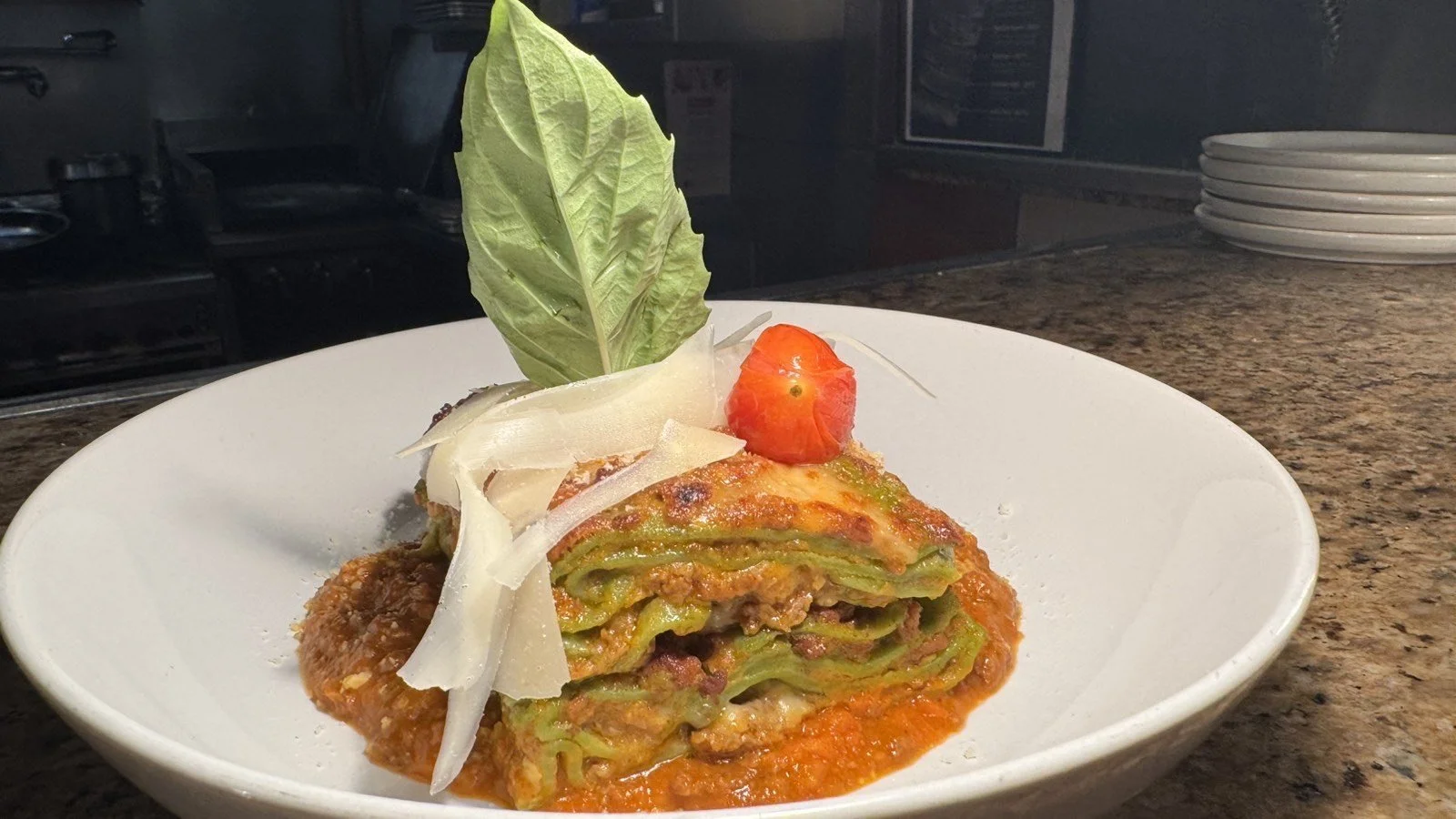 A serving of layered vegetable lasagna garnished with tomato, cheese, basil leaf, and onions in a white bowl on a kitchen countertop.
