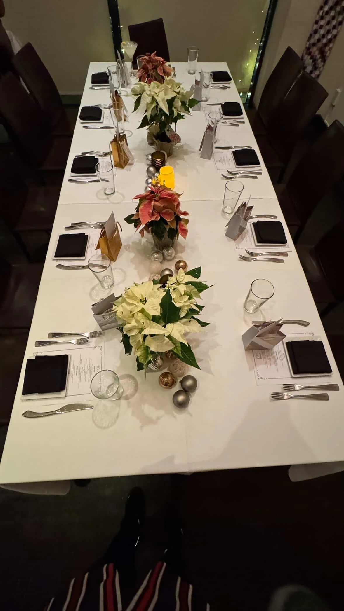 A rectangular dining table decorated for a holiday meal with poinsettia flowers in the center, Christmas ornaments, candles, and place settings that include black napkins, menus, silverware, and empty glasses. The table is set for eight people with f