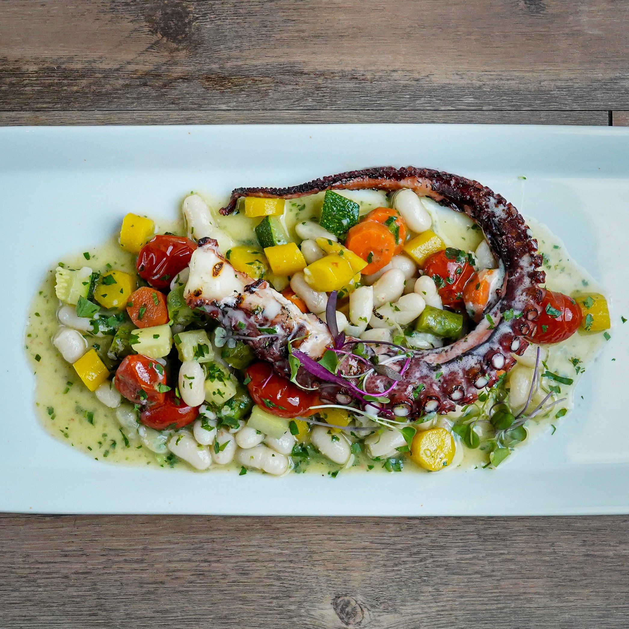 Grilled octopus tentacle on bed of white beans, roasted cherry tomatoes, and diced yellow and green vegetables, garnished with herbs and microgreens, served on a white rectangular plate.