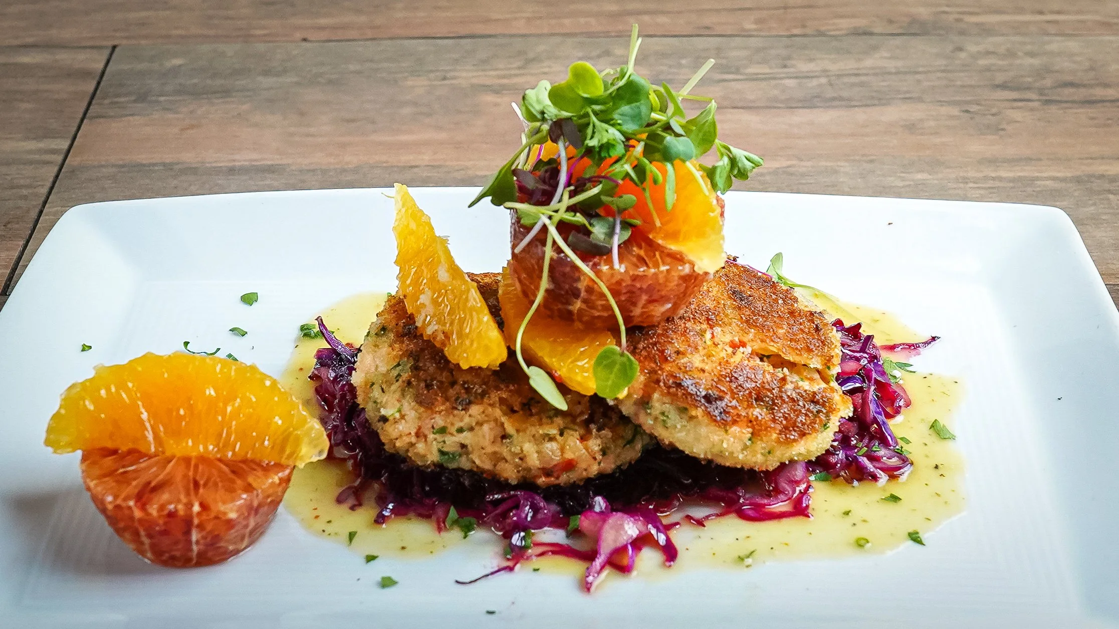 A white plate with cooked fish fillets garnished with microgreens, orange slices, and sauce, served on a bed of purple cabbage, with additional orange slices and sauce around it.