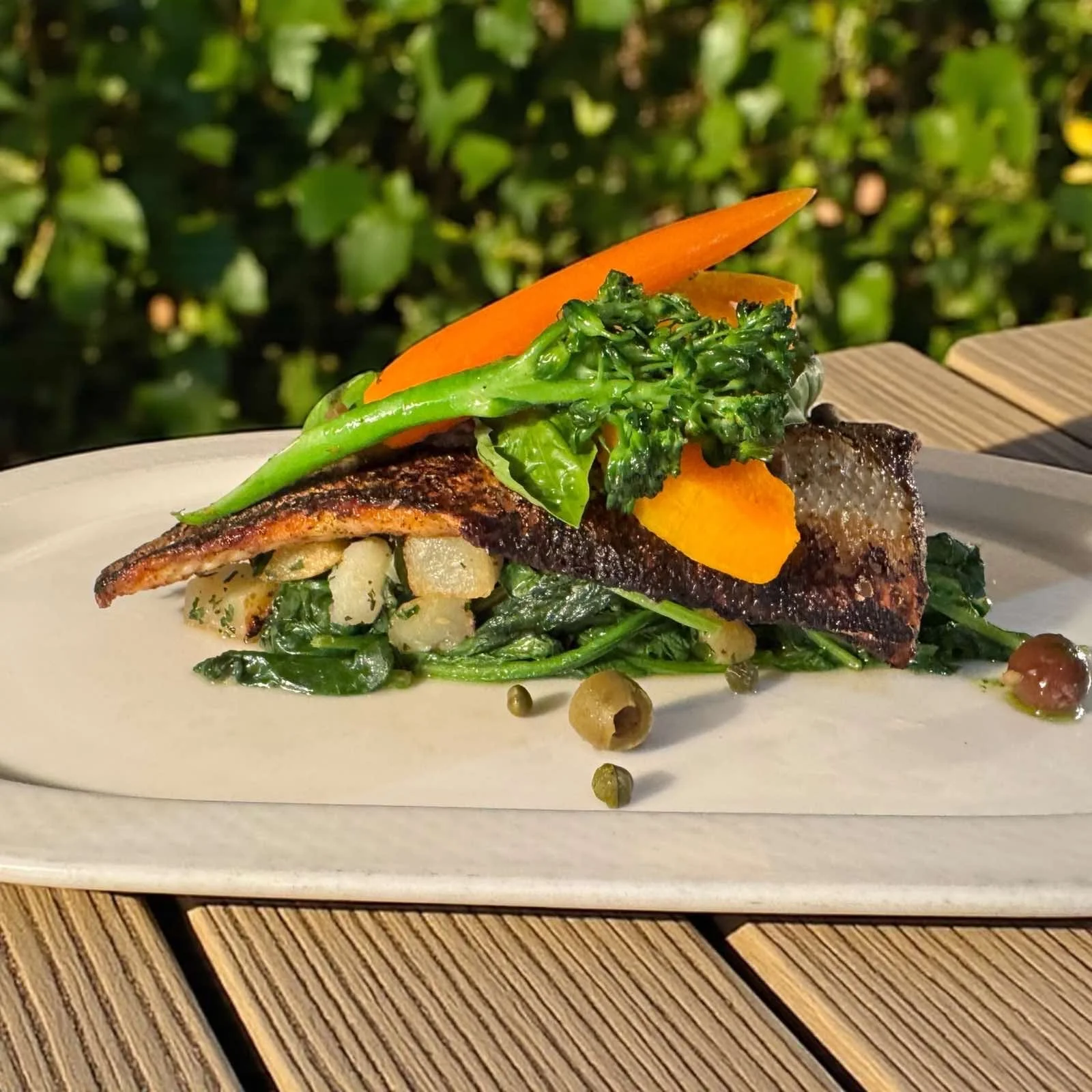A plated dish featuring a piece of grilled fish with crispy skin, topped with fresh broccoli and sliced carrots, served over sautéed greens and capers.