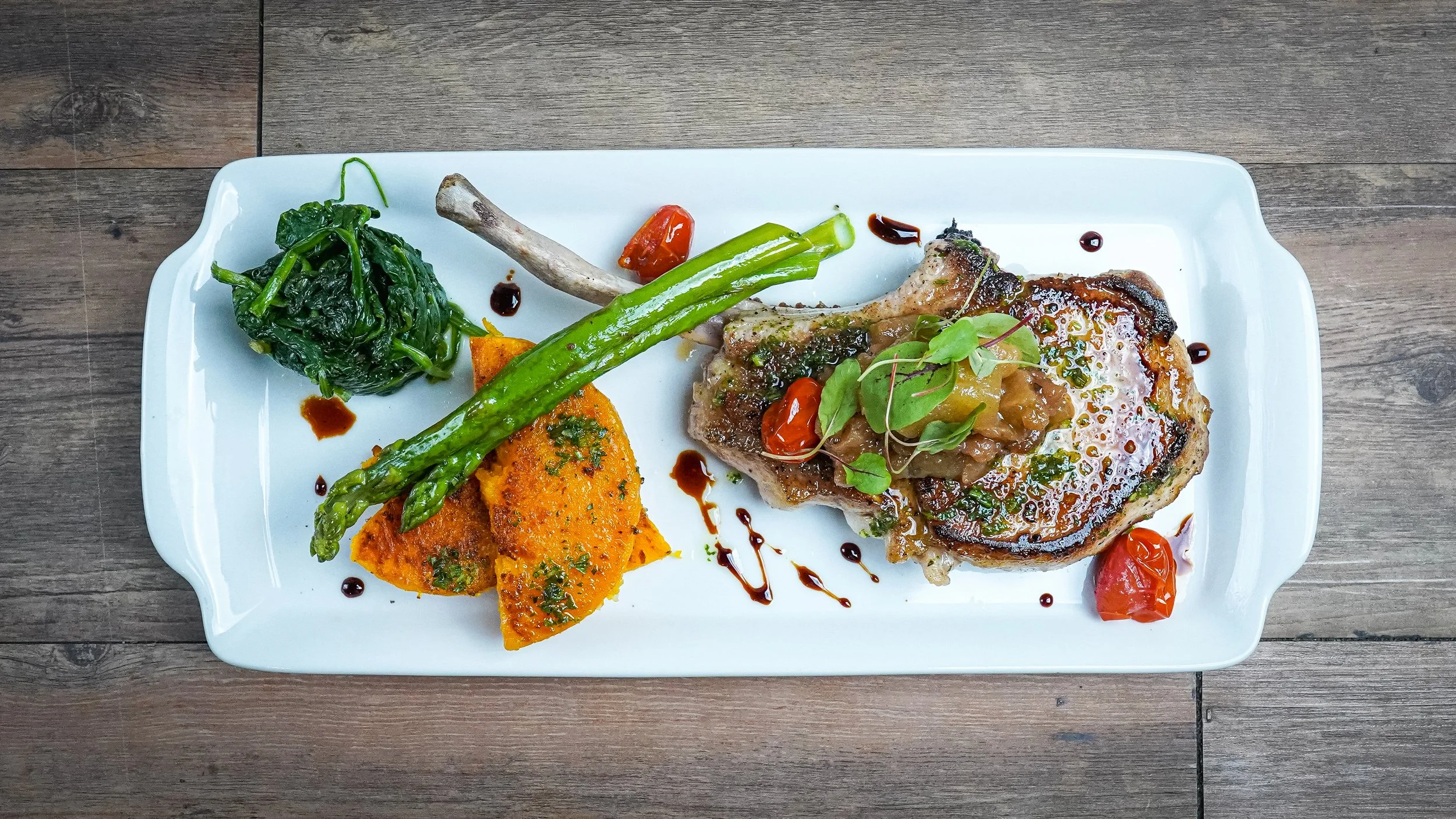 A plate of grilled steak with vegetables, including green beans, cherry tomatoes, and cooked spinach, garnished with microgreens and drizzled with sauce, served on a white platter.