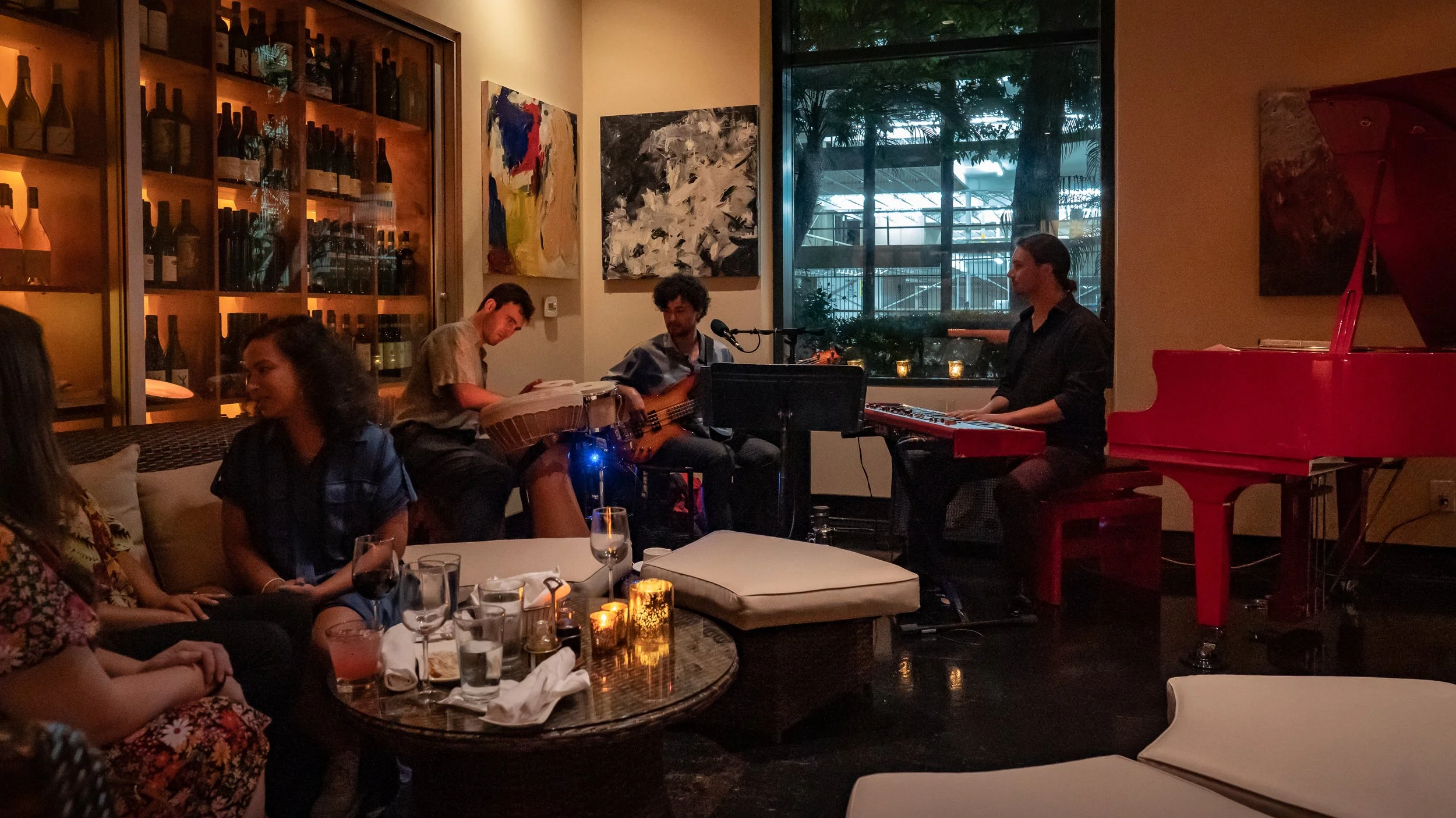 A group of four people performing live music in a cozy, dimly lit lounge with abstract paintings on the wall, a large window showing trees outside, and a bright red piano.