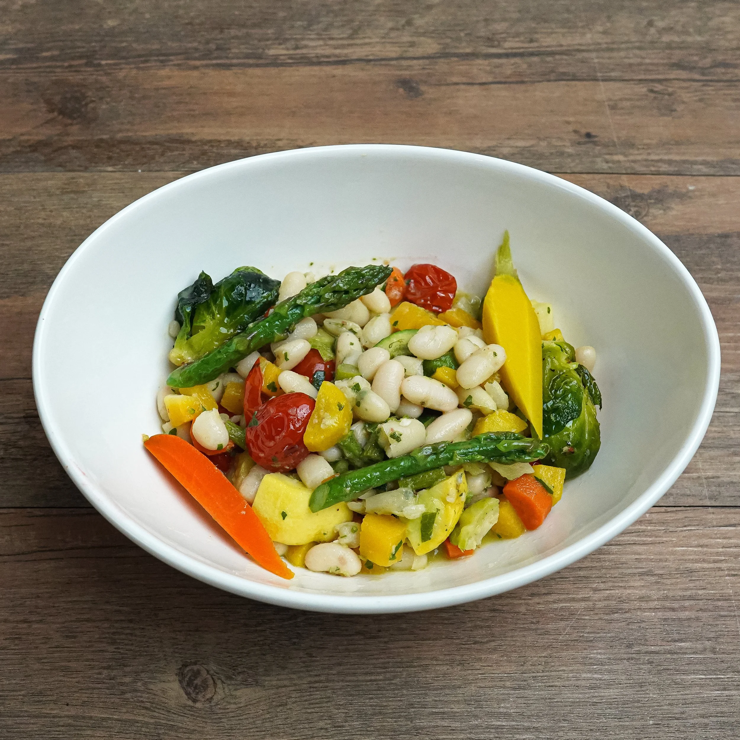 Bowl of mixed vegetable salad with white beans, cherry tomatoes, carrots, yellow squash, and asparagus on a wooden table.