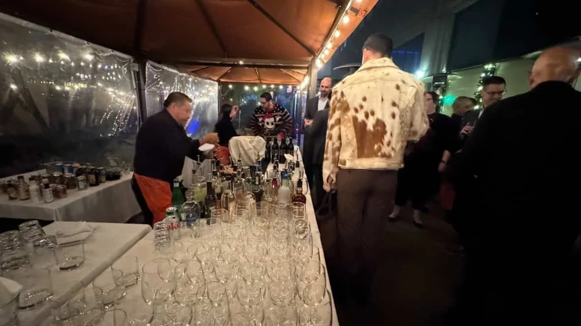 People mingling at a nighttime event with a bar setup, glasses on the table, and string lights overhead.