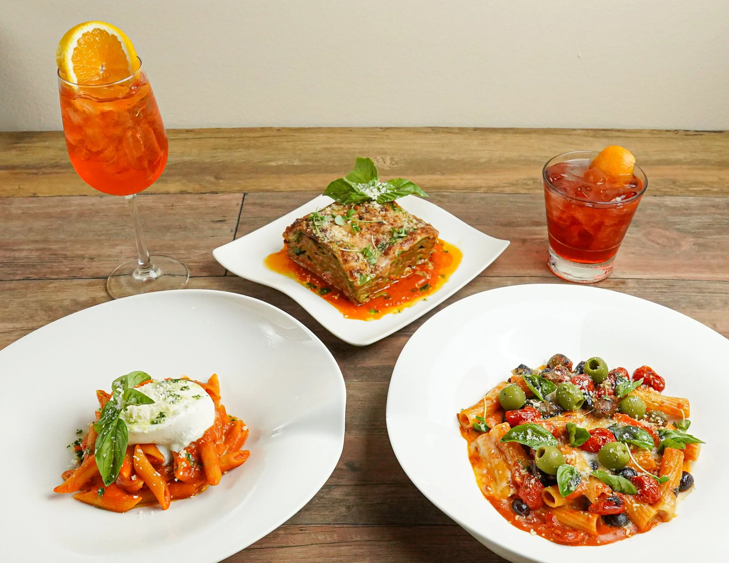 Three plates of Italian pasta, two cocktails with orange slices, and a dish of lasagna on a wooden table.