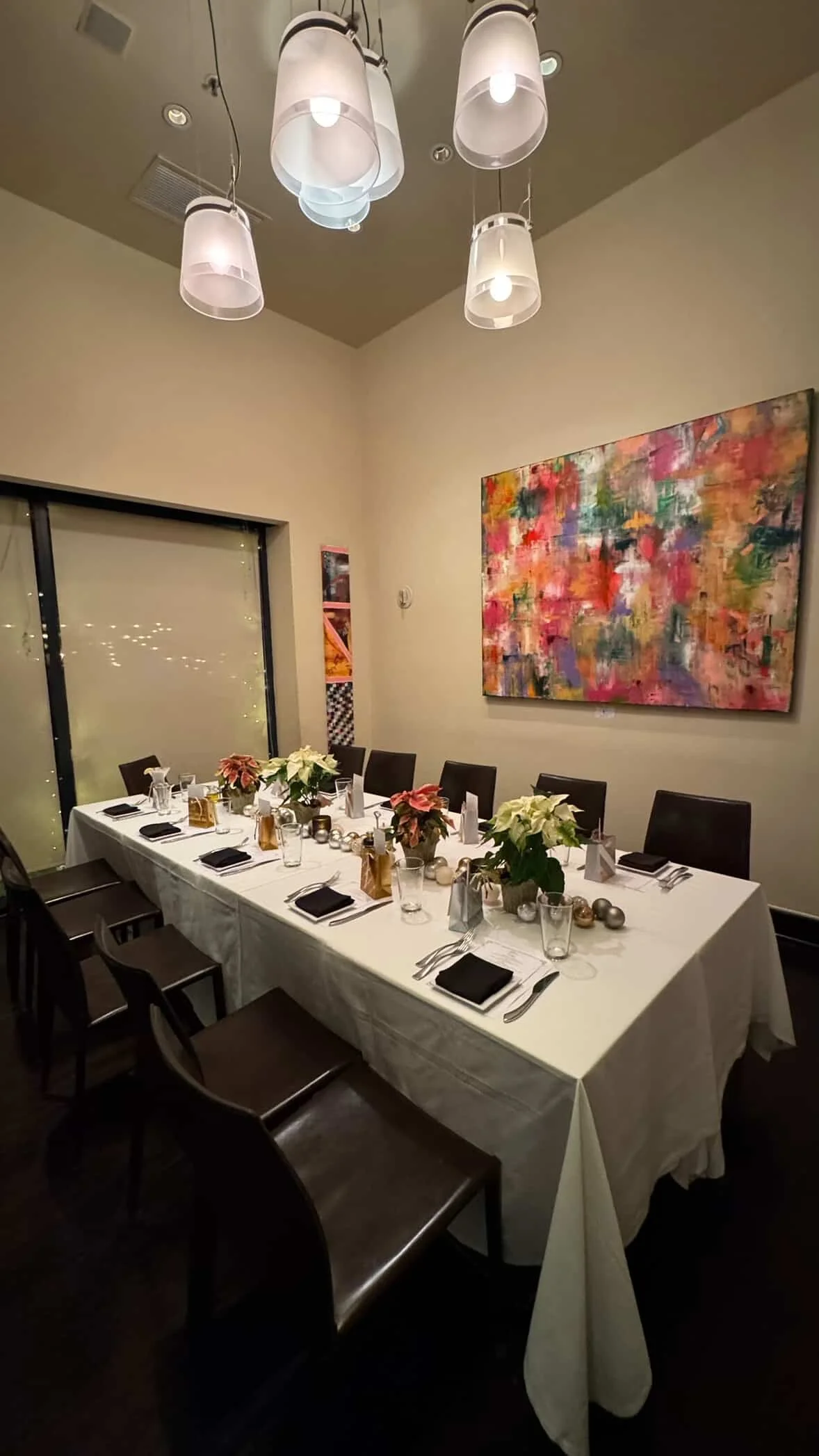 A dining table set for a celebration with black napkins, glassware, and Christmas-themed decorations, including poinsettia plants and silver ornaments, in a room with beige walls, abstract artwork, and modern pendant lighting.