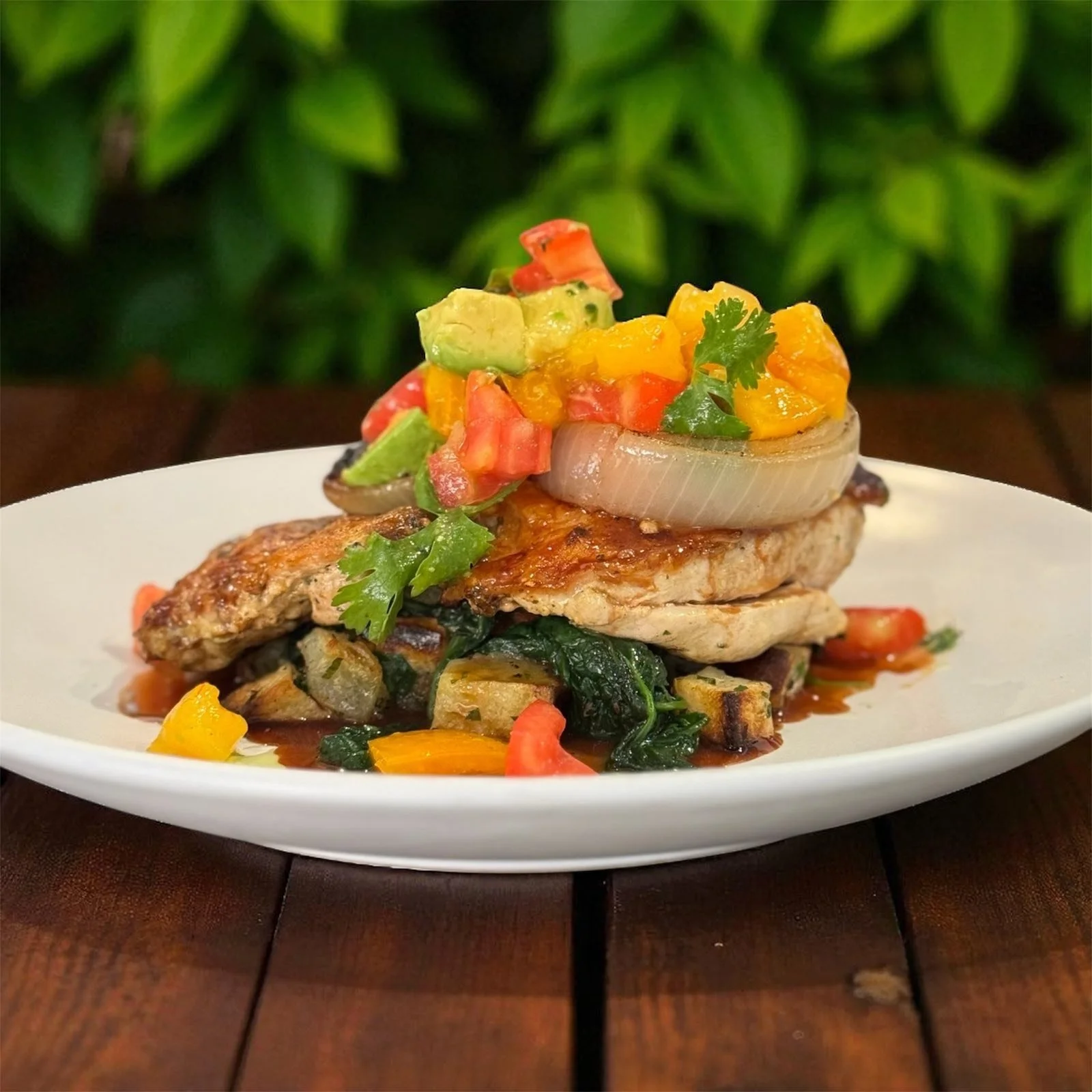 A plated dish featuring grilled chicken topped with chopped vegetables and an onion, garnished with cilantro, on a white oval plate with a dark wooden table and green leafy background.