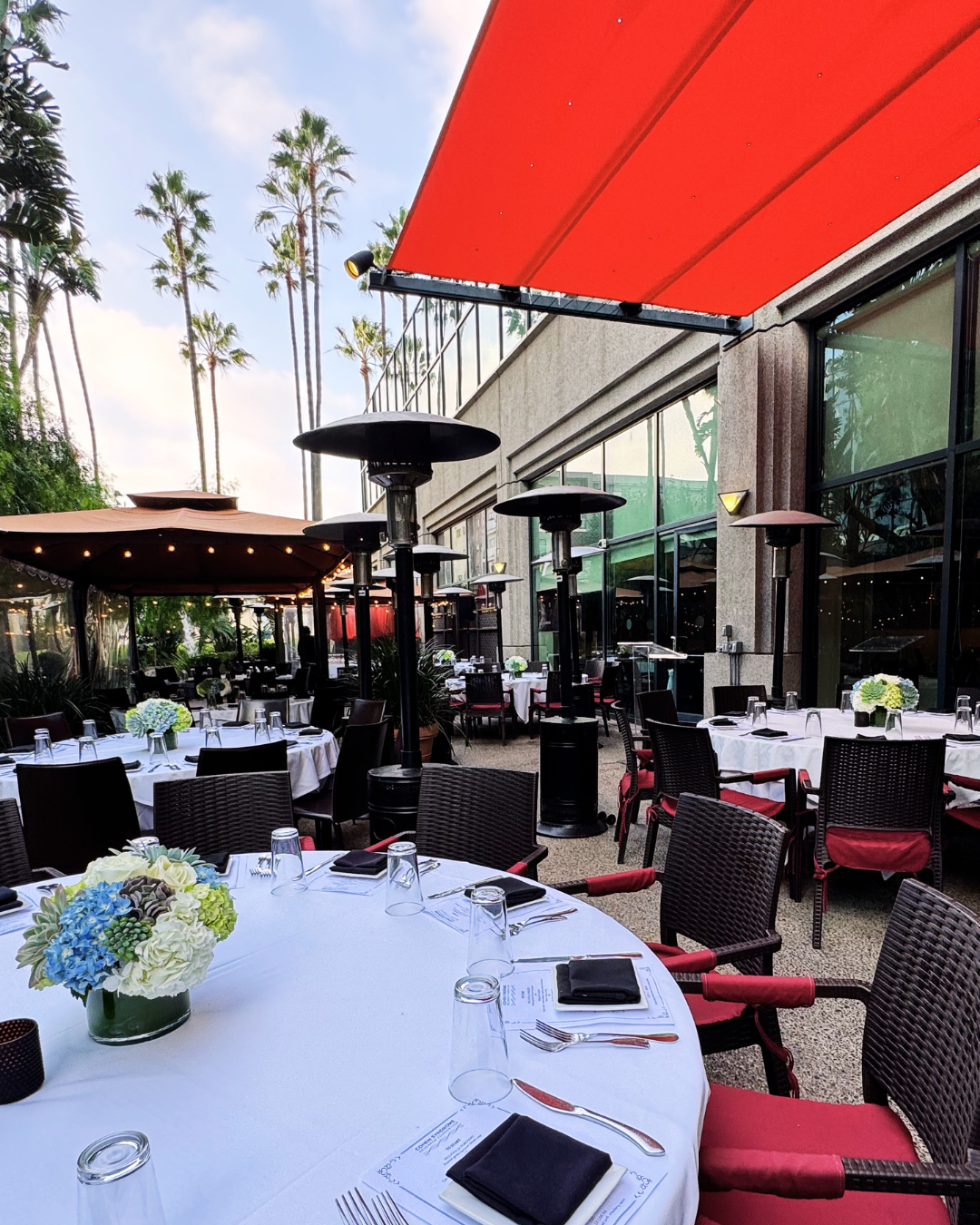 Outdoor restaurant patio with round tables covered in white tablecloths, decorated with flower centerpieces, set with glasses, utensils, and black napkins. There are black wicker chairs with red cushions, patio heaters, and a red awning overhead. Tall palm trees are visible in the background under a partly cloudy sky.