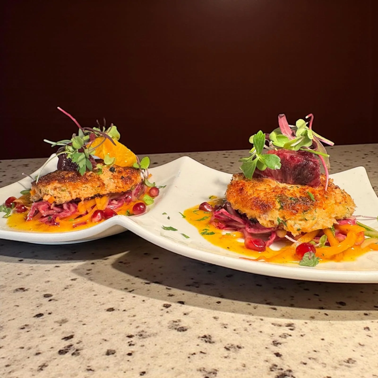 Two plates of breaded fish fillets served over shredded vegetables, garnished with microgreens and orange slices, with a sauce drizzled underneath, on a speckled countertop.