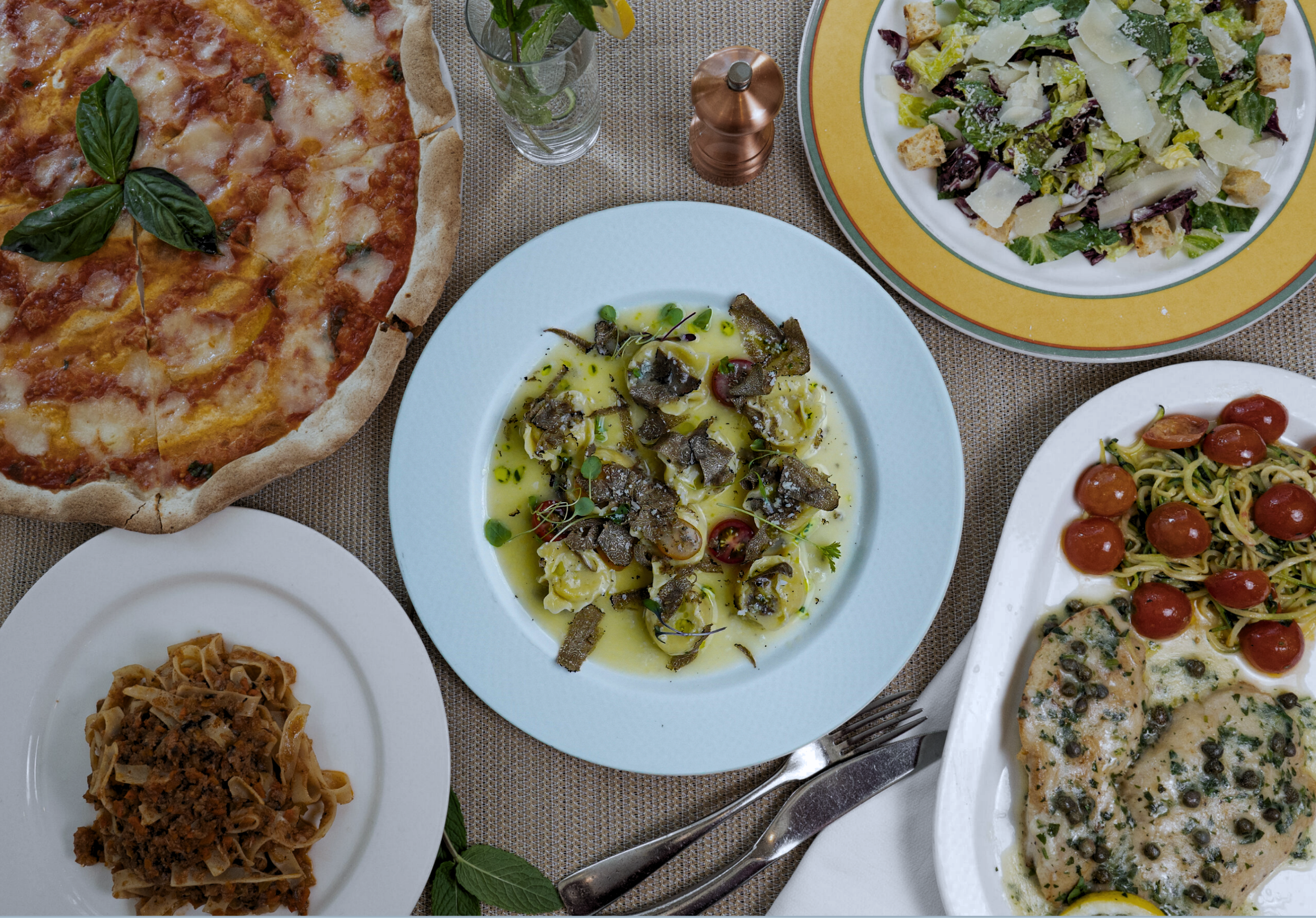 A spread of Italian dishes and a salad on a table, including a pizza with basil, a plate of pasta with meat sauce, a plate of ravioli with sauce, a mixed green salad with croutons and cheese, a plate of pasta with cherry tomatoes and chicken with capers, and a plate of pasta with truffles.