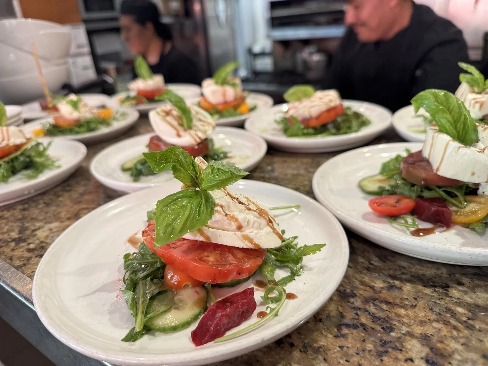 Plates of Caprese salad with sliced tomatoes, fresh mozzarella, basil leaves, and balsamic glaze garnished with a cherry tomato and mixed greens on a granite countertop, in a restaurant kitchen.