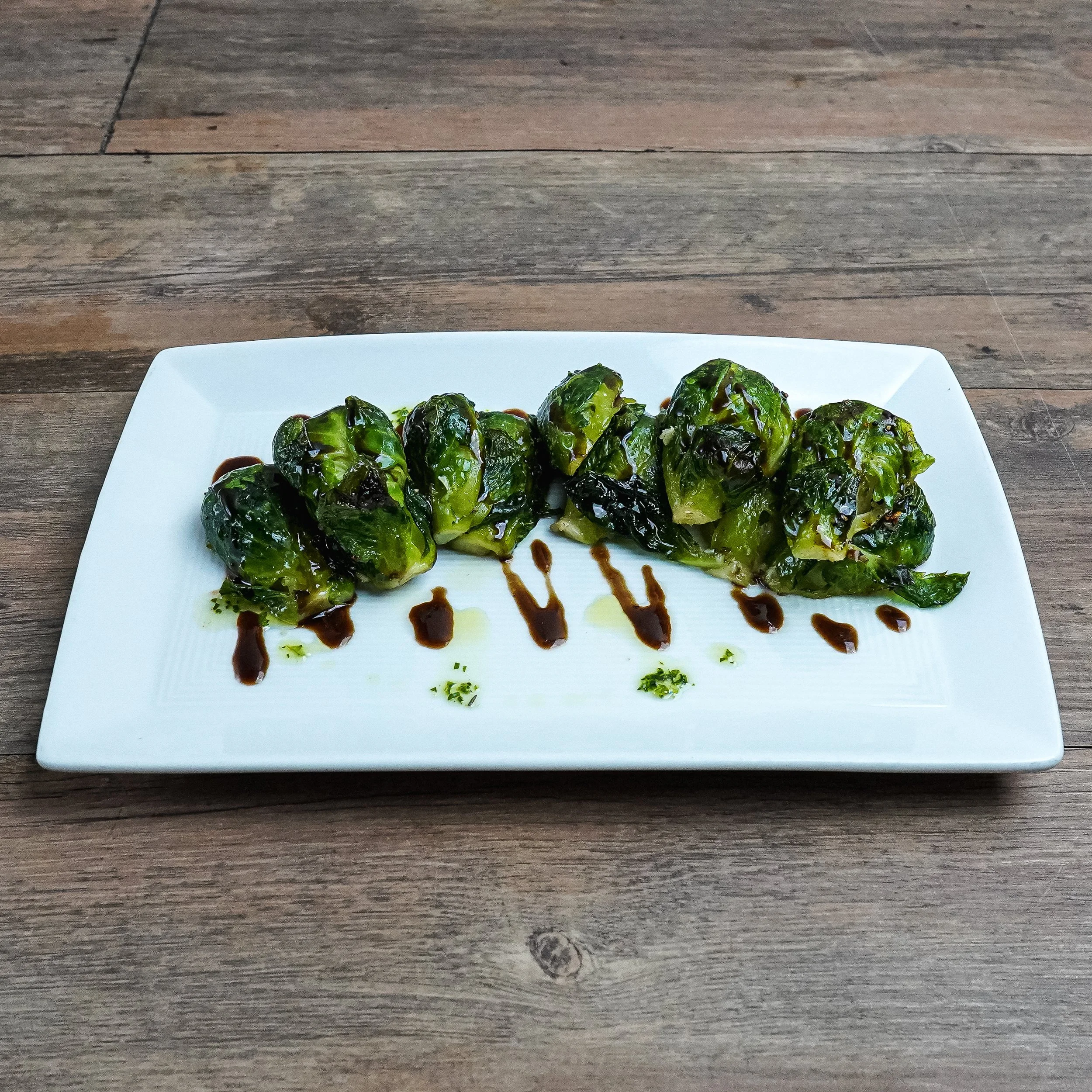 Six cooked Brussels sprouts drizzled with dark sauce and olive oil on a white rectangular plate, placed on a wooden table.