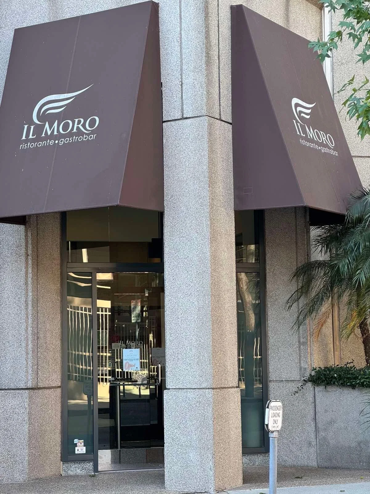 The exterior of a restaurant called Il Moro with two brown awnings displaying the restaurant's logo and name. The entrance is a glass door, and next to it is a sign that says 'Passenger Loading Only, Shuttle'.