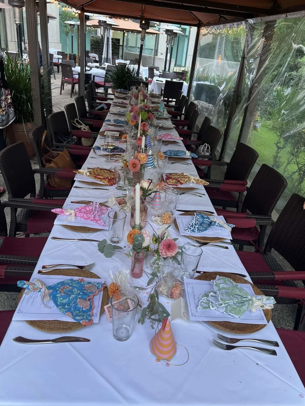 Long dining table set for a celebration with colorful party hats, flowers, and place settings, under a canopy on an outdoor patio.