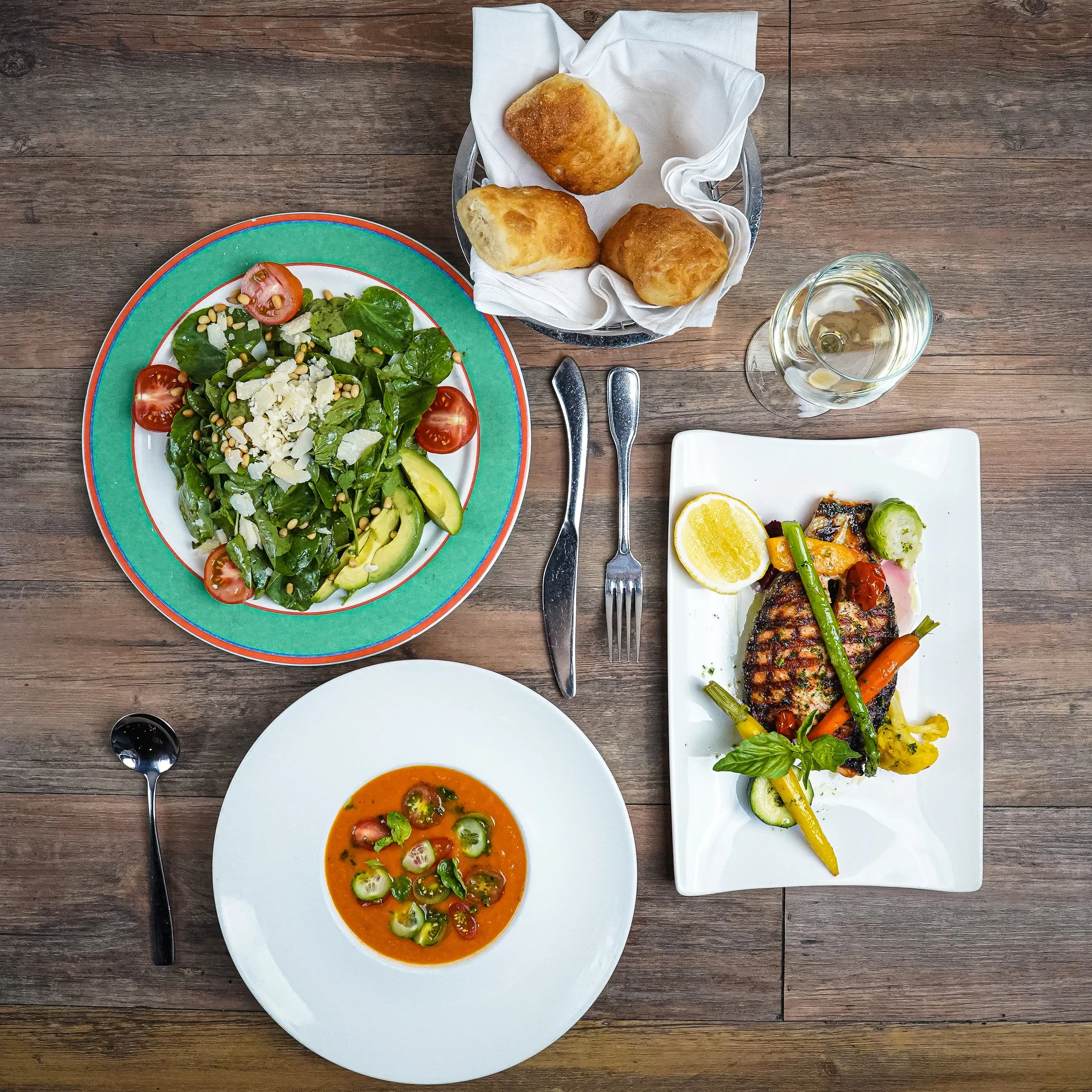A wooden table with a colorful salad, a bowl of tomato soup, a plate of grilled fish with vegetables, a basket of bread rolls, and a glass of white wine.