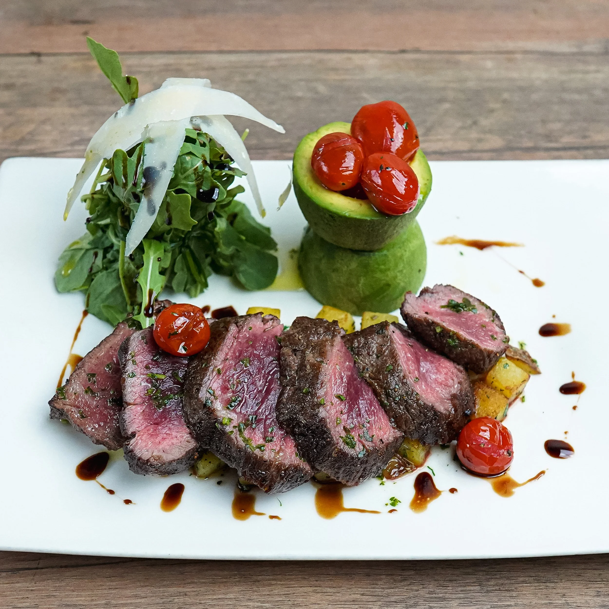Sliced seared steak served with greens, cherry tomatoes, and avocado with cherries inside, drizzled with balsamic vinegar on a white rectangular plate.