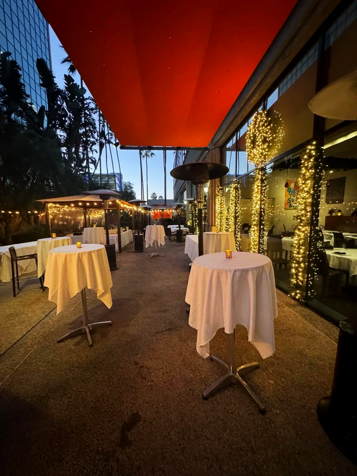 An outdoor restaurant patio at dusk with several round tables covered in white tablecloths and small candles. The space is decorated with string lights and holiday decorations, including Christmas trees. There are large patio heaters, umbrella-covered tables, and a large red awning overhead. Tall palm trees can be seen in the background.