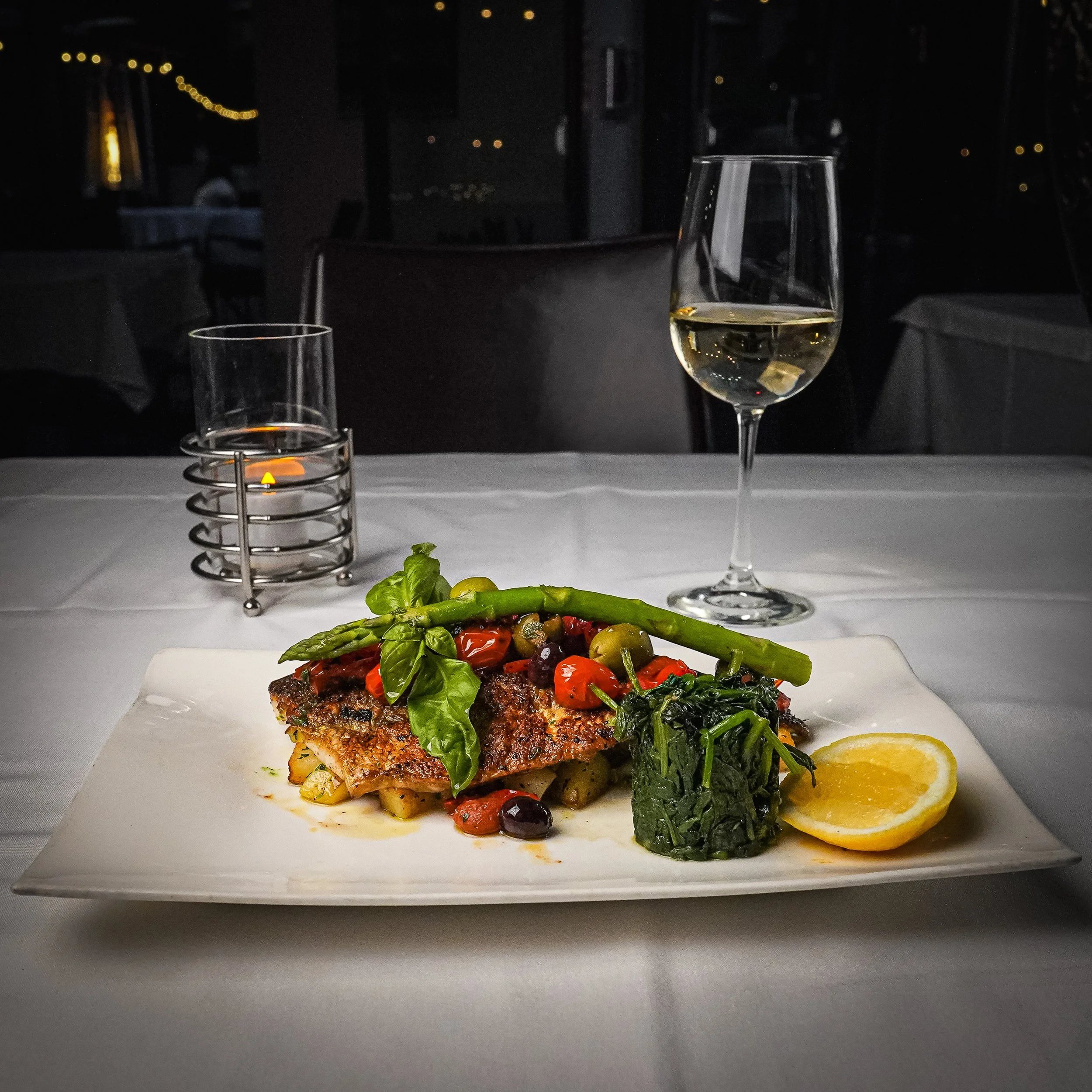 A plated serving of grilled fish with vegetables, a lemon wedge, a glass of white wine, and a lit candle on a restaurant table at night.